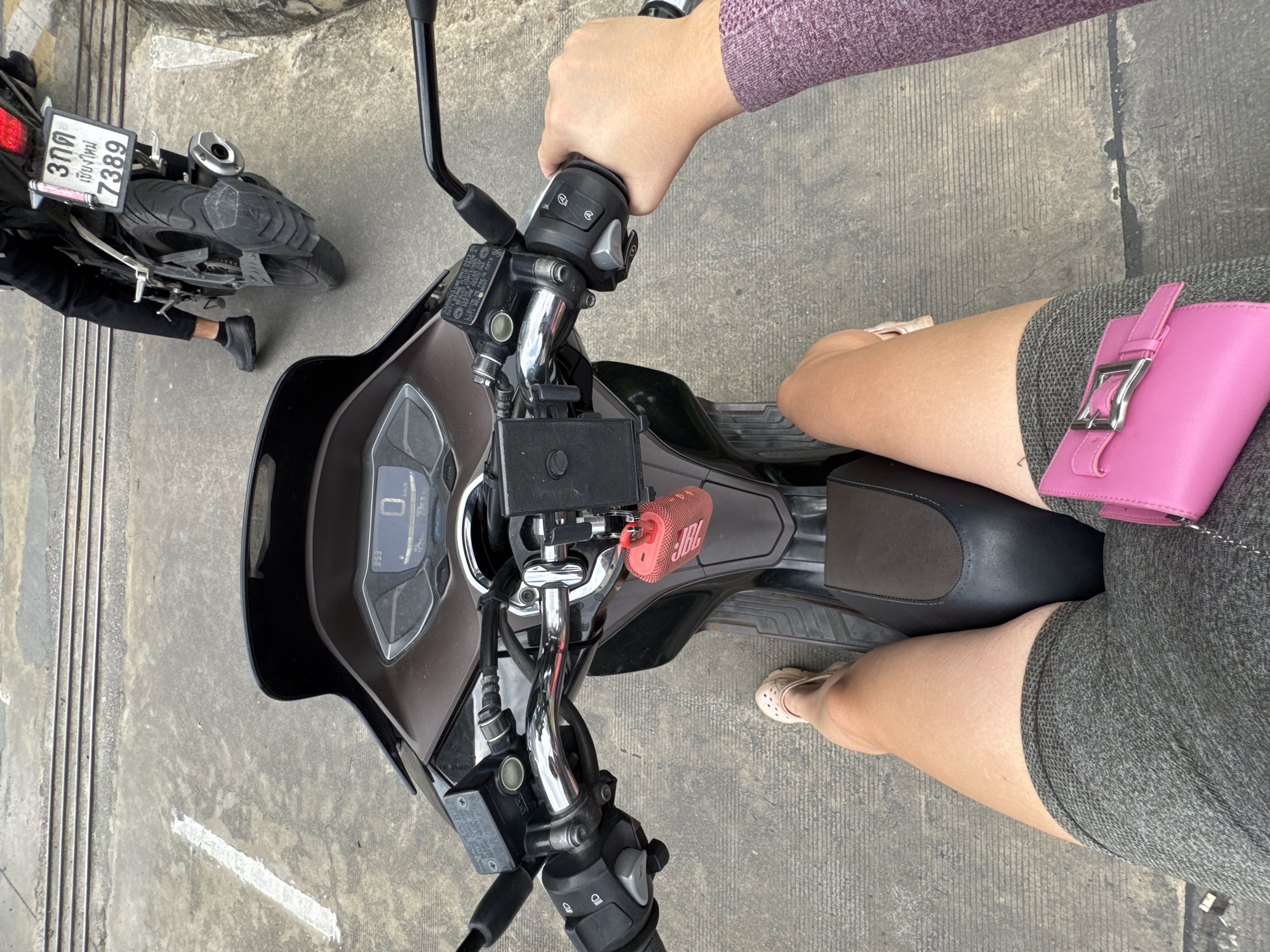 This media file is a top-down shot of a person sitting on a scooter, likely in Nong Pa Khrang, Thailand. The scooter's dashboard is visible, showing a digital display with a speed reading of "0". Attached to the scooter is a small, red JBL speaker. The rider is wearing a grey skirt and pink sandals. To the left of the scooter, another motorcycle is partially visible, with a license plate displaying "3กต เชียงใหม่ 7389". The ground appears to be concrete, with some lines and debris. The scene suggests the person is about to start riding or has just stopped. The lighting indicates it is daytime.