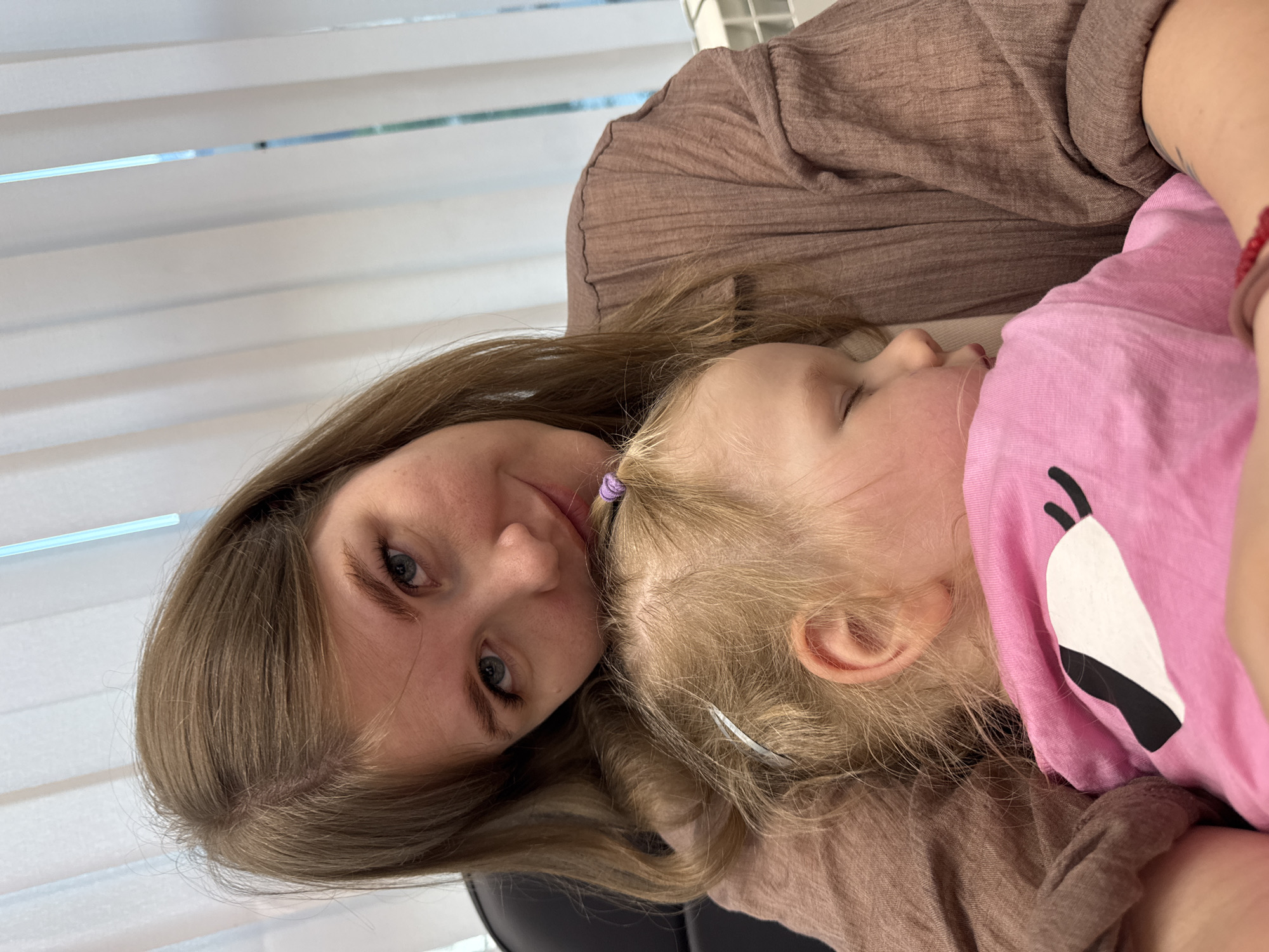 A woman with light brown hair and blue eyes is holding a blonde-haired child wearing a pink shirt with a bird design. The child is asleep. The woman is looking at the camera with a slight smile. The background appears to be a room with white blinds. The location in the city of Perm, Russia, cannot be determined based on the image.