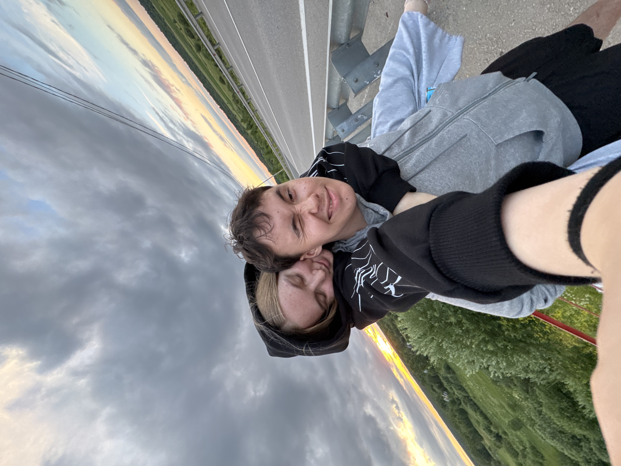 Two young people are captured in a selfie, seemingly on a bridge or overpass, with a scenic rural landscape and a road visible in the background. The person in front, a male, is looking at the camera and winking, with his mouth slightly open. He is wearing a grey hooded sweatshirt. Behind him, a female is hugging him from behind, with her eyes closed and head resting on his shoulder. She is wearing a black hooded sweatshirt with white graphic detailing. The sky above is dramatic with dark clouds and patches of sunset light. The road in the background appears to be a highway with white lane markings, bordered by a guardrail. Below the road, a green, hilly landscape with trees is visible. The image was taken at sunset, as indicated by the warm light in the sky. The location is identified as Ryabinino, Russia.