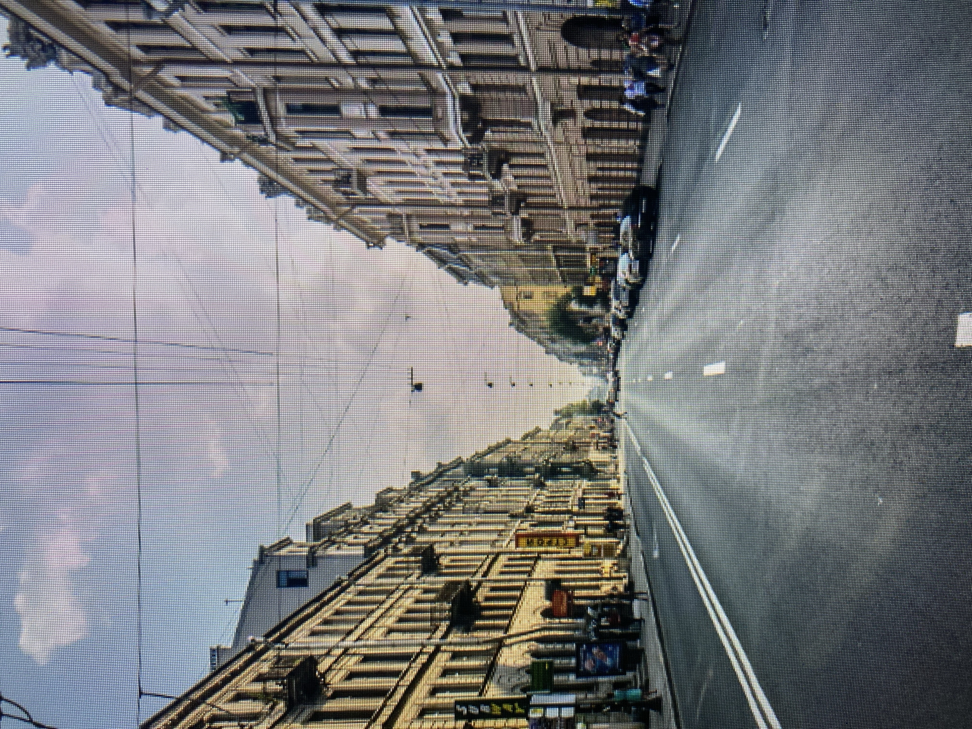 The image depicts a wide city street lined with tall, multi-story buildings on both sides. The buildings appear to be old, with classical architectural details, and are painted in light colors. There are balconies on some of the buildings. The street is paved and has double white lines marking lanes. Several cars are parked along the side of the road on the right. Pedestrians are visible walking on the sidewalks on both sides of the street. Overhead, there are numerous electrical wires crisscrossing the sky, attached to poles and buildings. The sky is partially cloudy with a light blue hue. The scene suggests a day in an urban environment. The provided context of Naberezhnye Chelny, Russia, is not evident from visual cues within the image itself, as there are no discernible landmarks, signs, or architectural styles that specifically identify this location.