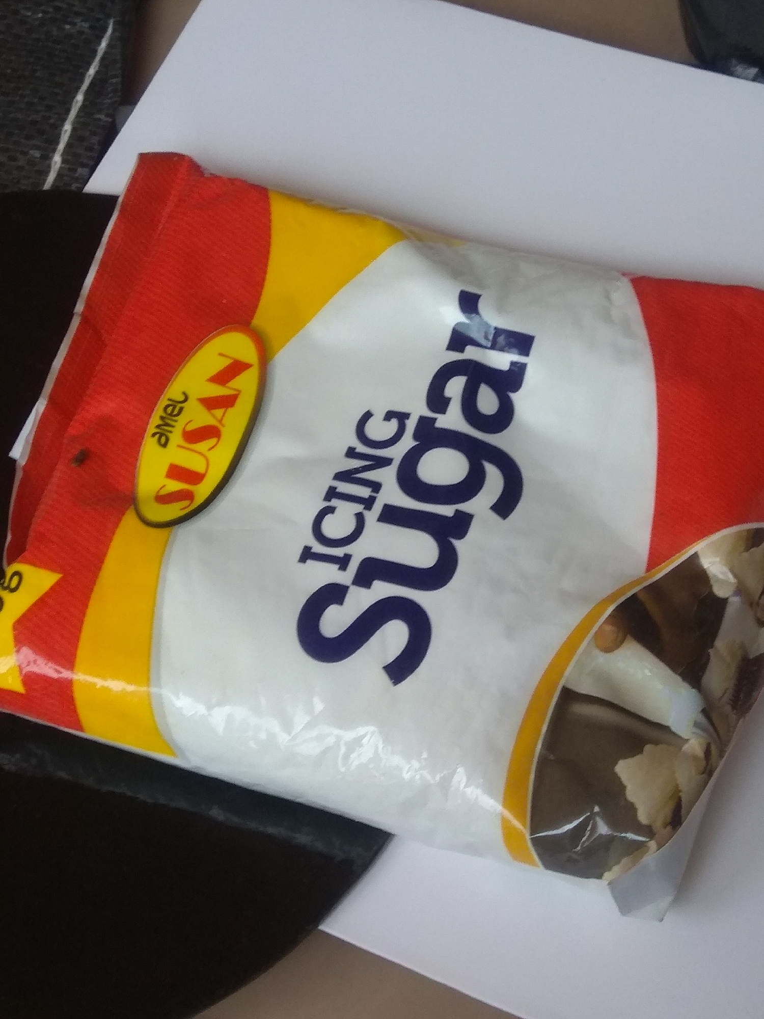 A bag of "Amel Susan" brand icing sugar is laid on a white surface, partially overlapping a dark surface. The bag is primarily white with red and yellow accents. The text "ICING Sugar" is prominently displayed in dark blue lettering, with "Amel SUSAN" in a yellow oval on the left side of the bag. The sugar inside the bag is visible through a translucent section on the right, appearing as a light-colored granular substance. The overall lighting is moderate, casting soft shadows. The image is taken from a slightly elevated angle. There are no people or animals visible in the image. The scene appears to be indoors, with no indication of a specific location in Jalingo, Nigeria.