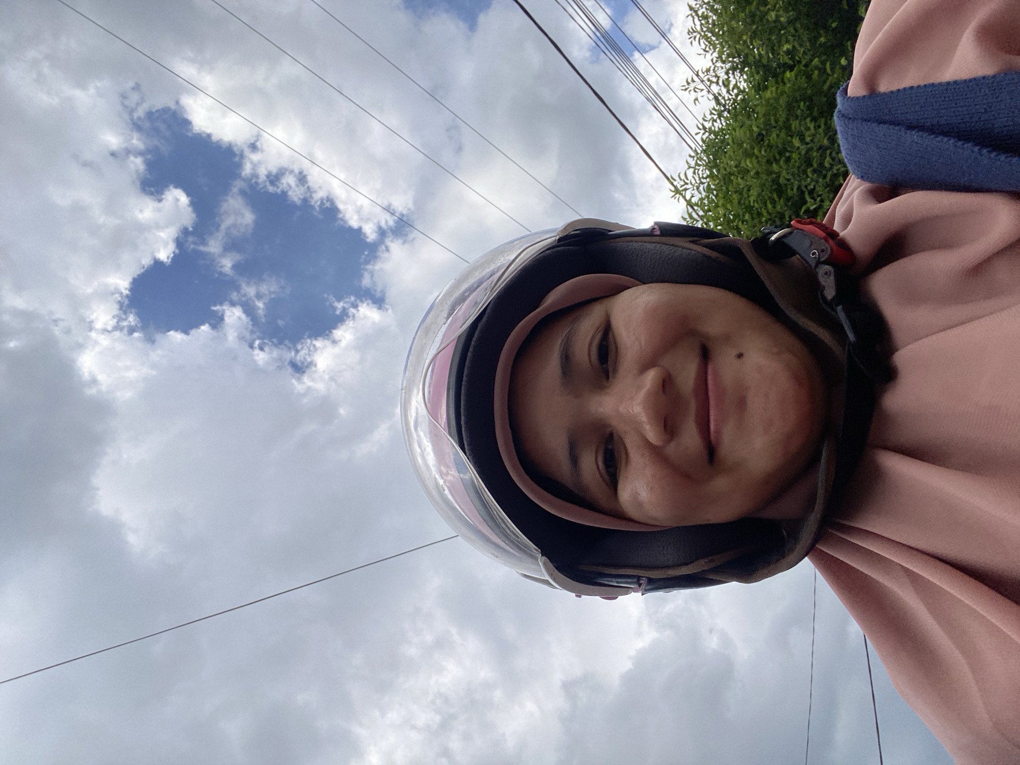 A young Indonesian woman is taking a selfie while wearing a helmet and a pink hijab. She has a bright smile and is looking directly at the camera. The background shows a cloudy sky with some visible power lines. Behind her, green trees are partially visible. The overall setting suggests an outdoor environment, possibly a street or roadside in Banjarbaru, Indonesia.