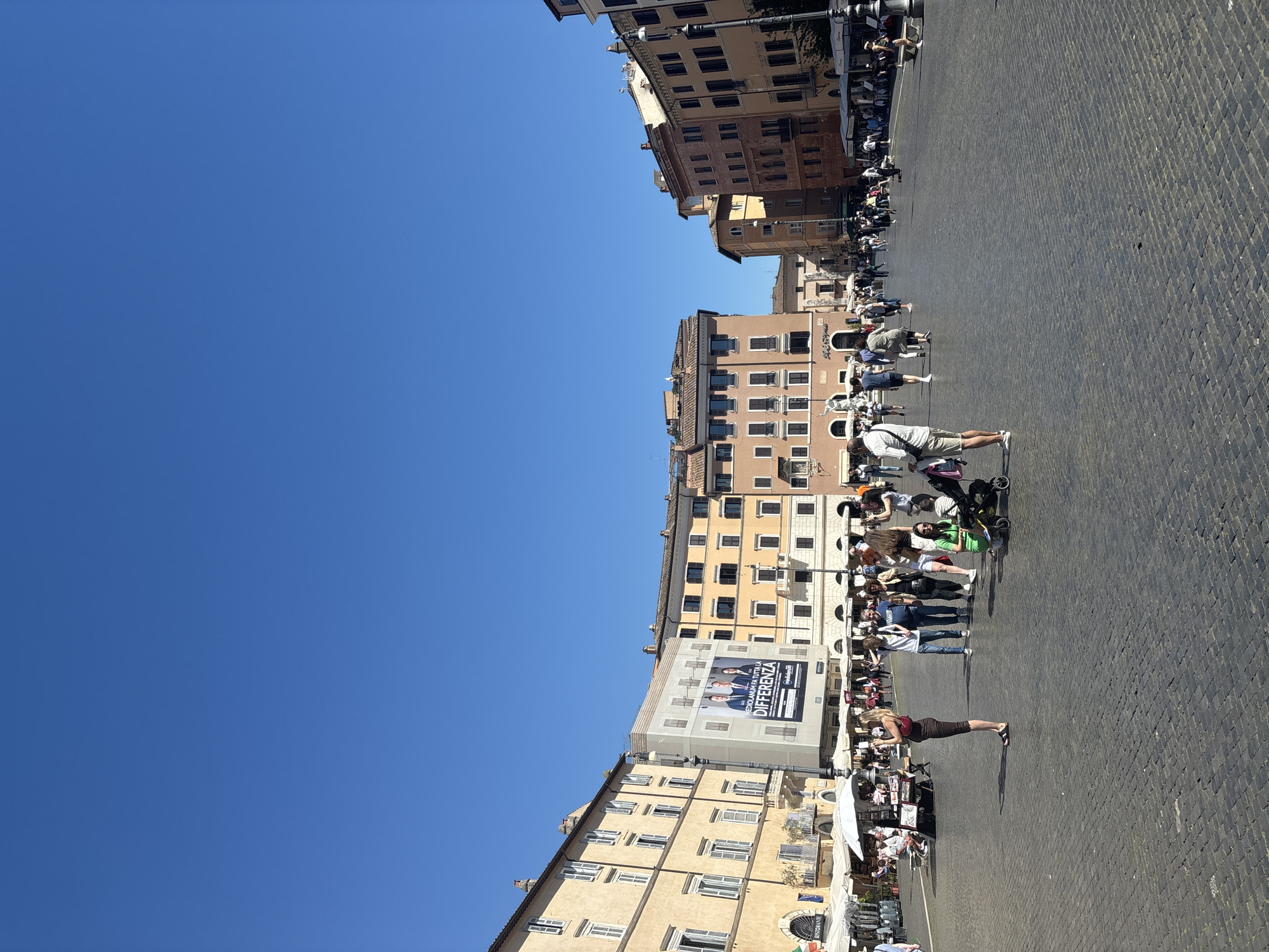 The image depicts a sunlit, cobblestone piazza in Rome, Italy, with a clear blue sky above. Buildings line the square, including several with tan facades and numerous windows. A large political advertisement banner is visible on one building. A group of people is gathered, mostly standing and some pushing strollers. Several people are interacting with displays or tables on the square. The ground is a cobblestone surface.