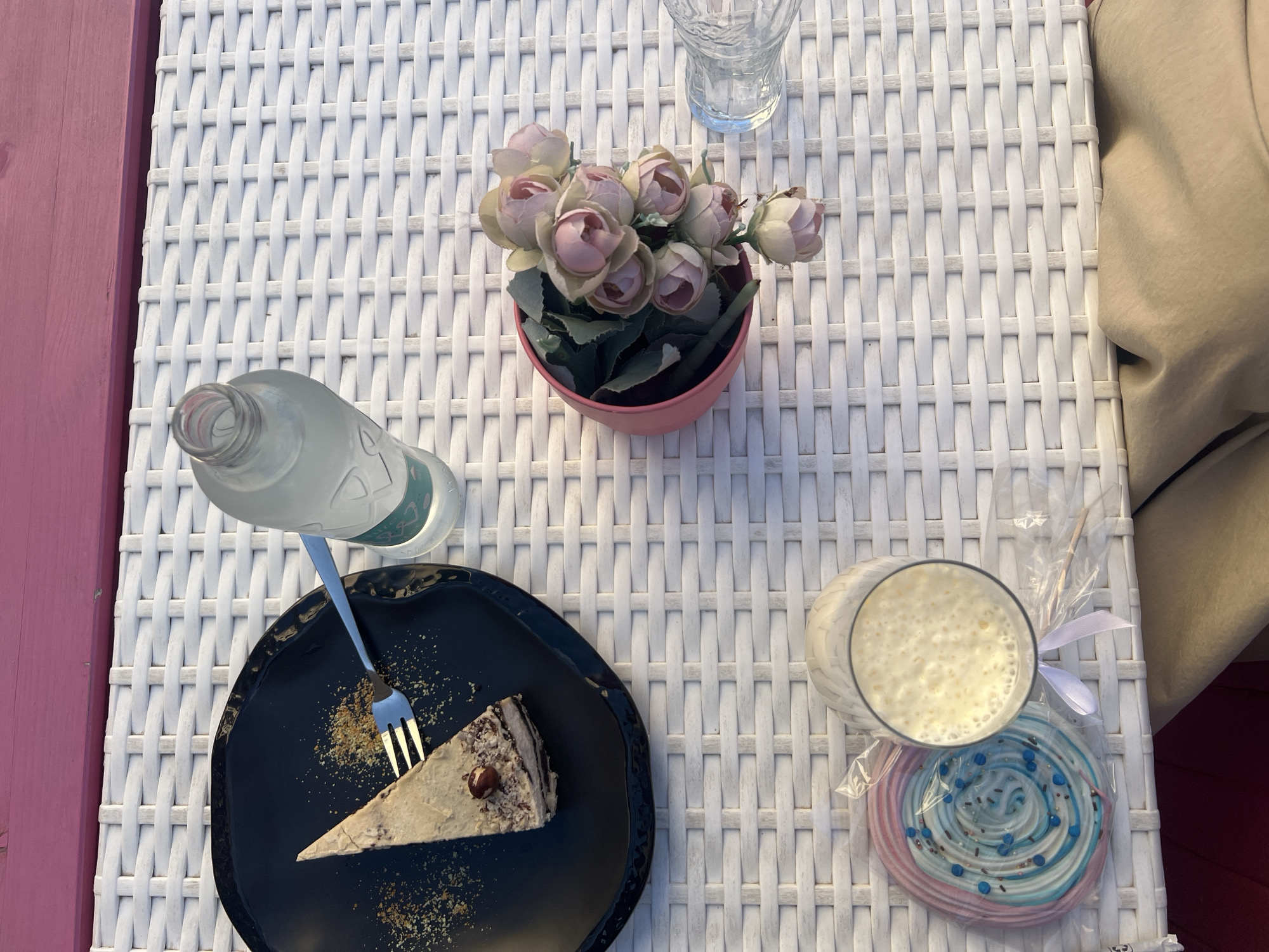The image depicts a table setting in what appears to be an outdoor café, possibly in Plovdiv, Bulgaria. The table is made of white wicker. On the table, from left to right, is a glass bottle, a black plate with a slice of cake and a fork resting on it, a pink pot of artificial pink roses, a clear drinking glass, a glass of milk, and a packaged blue and pink lollipop. A woman in a tan jacket is sitting on the right. The edge of a pink wall or bench is visible to the left.
