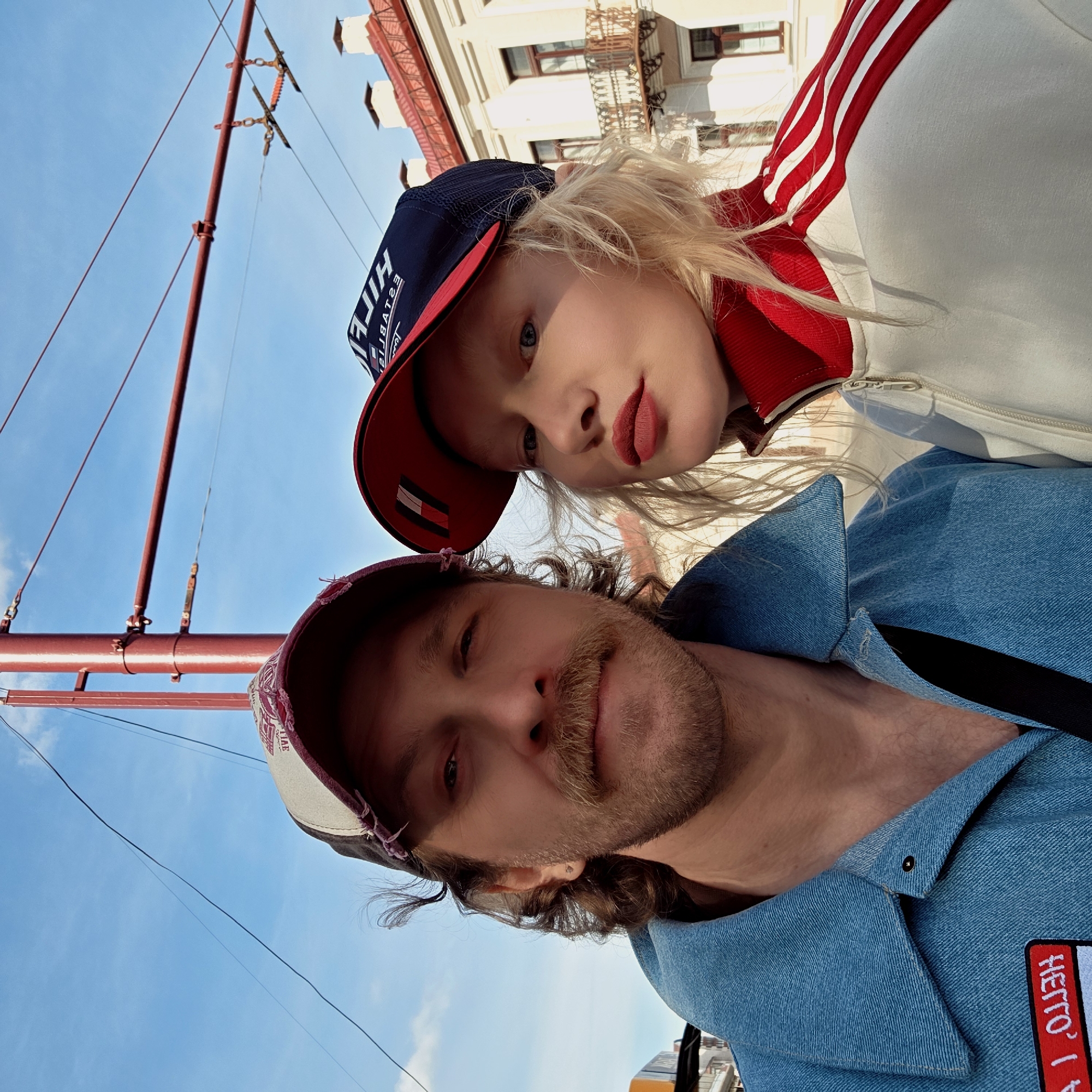 A low-angle selfie captures two individuals, a man and a woman, against a bright blue sky in an urban setting, likely Hrodna, Belarus. The image appears to be rotated approximately 90 degrees counter-clockwise.
In the foreground, a woman with blonde hair wears a dark blue baseball cap with a red brim, featuring the mirrored text "HILFIGER" and "ESTABLISHED". She is dressed in a white jacket with red and white stripes along the shoulder and arm. Her face is turned slightly towards the camera, showcasing striking red lipstick and blue eyes. To her left, a man with a beard and mustache looks directly at the camera. He wears a light blue denim jacket, with a red and white patch bearing the mirrored text "HETTO?". He also has a baseball cap on, which appears distressed or worn, with unclear markings.
The background reveals a clear, bright blue sky with sparse white clouds, indicating sunny weather. Red-painted metal poles and thin overhead wires, consistent with tram lines or utility infrastructure, crisscross the upper part of the frame. Behind the woman, part of an ornate, light-colored building with windows and reddish architectural elements is visible, suggesting classic European architecture. At the very bottom edge of the image, a glimpse of a yellow-orange vehicle, possibly a tram or bus, can be seen. Both individuals maintain relatively neutral expressions as they pose for the photo.