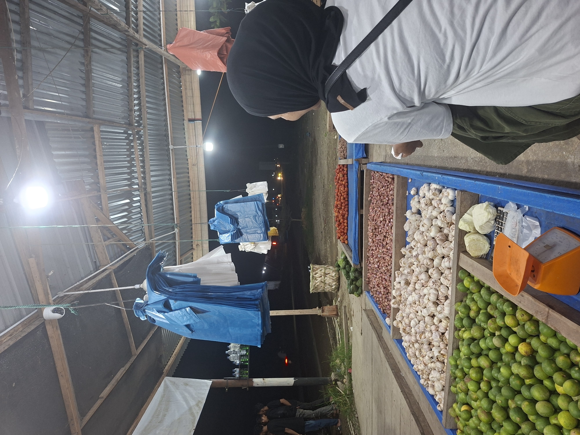 A person wearing a black hijab and white shirt stands at a stall filled with produce. The stall contains, from left to right: tomatoes, onions, garlic, cabbages, and limes. There is an orange scale at the edge of the stall. In the background, under a corrugated metal roof, blue tarps hang. In the foreground, people are standing near the edge of the structure, beneath the hanging tarps. The location appears to be Keudeteupinpunti, Indonesia. It is nighttime.