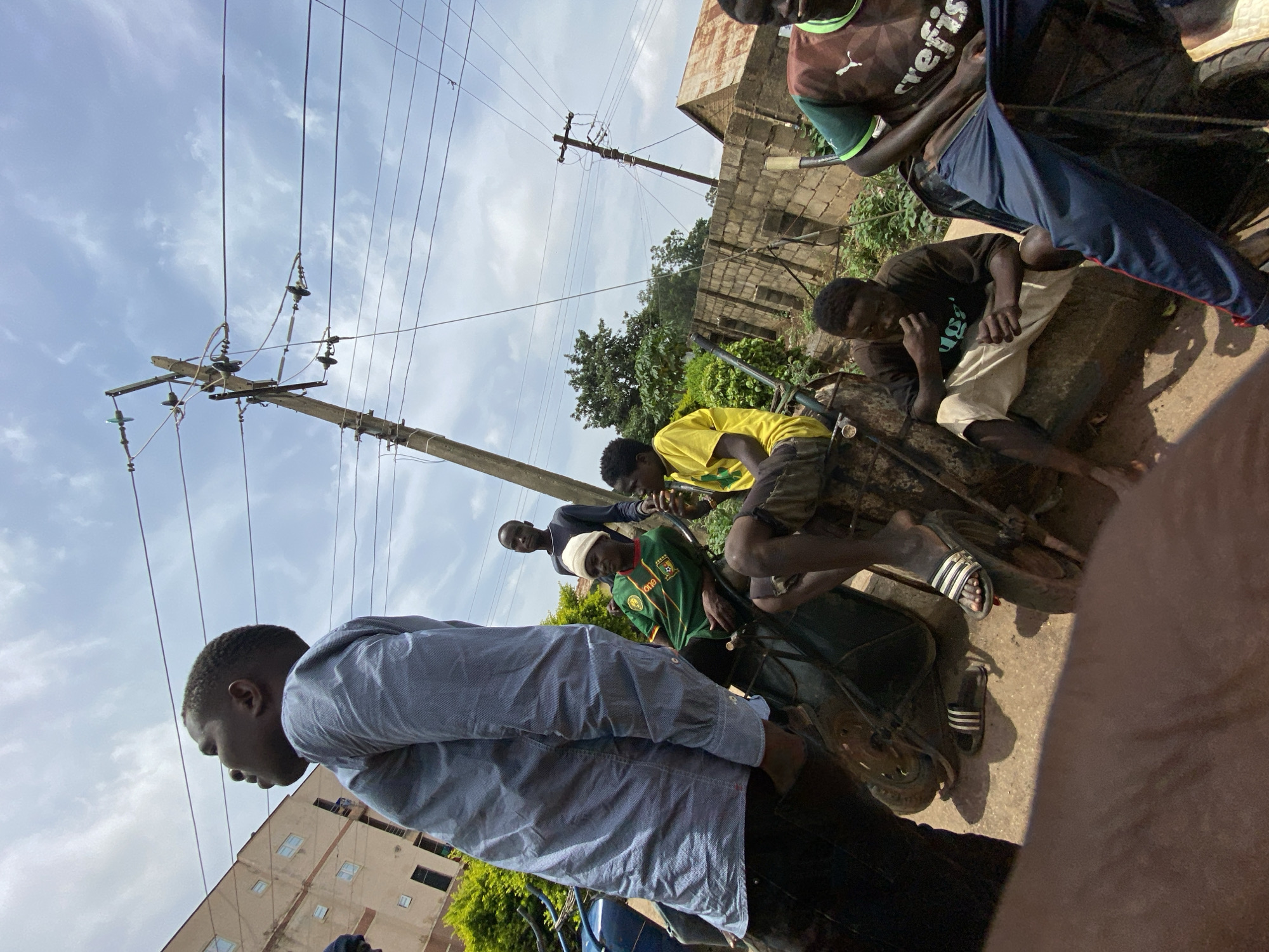 This image captures a street scene in Bukuru, Nigeria, with several young men gathered. In the foreground, a man wearing a light blue, patterned button-down shirt and dark pants is partially visible, looking upwards. To his right, a group of men are seated on or around wheelbarrows. One man wears a green and yellow football jersey with the Cameroon crest and a white beanie. Another man in a yellow t-shirt is visible, and a third in a dark t-shirt is eating. A fourth man in a brown and green t-shirt is seated on a wheelbarrow to the right. In the background, there are buildings, trees, and utility poles with numerous wires crisscrossing against a cloudy sky. The ground appears to be dirt or gravel. The overall impression is a casual gathering in an urban environment.