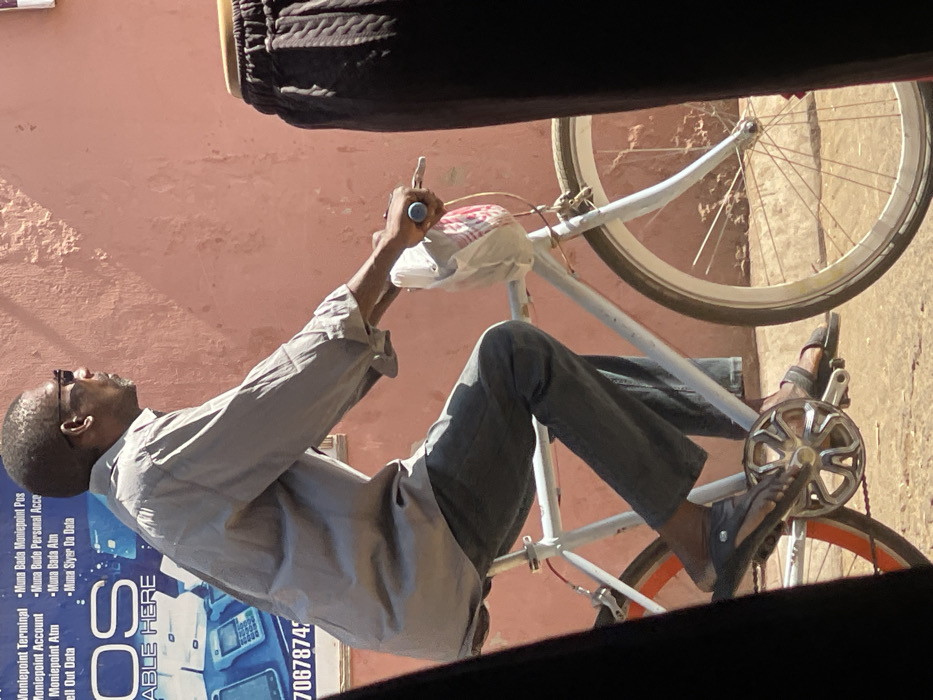 An adult male, dressed in a light grey long-sleeved shirt and dark grey trousers, rides a light-colored bicycle in Samaru, Nigeria. He wears dark sunglasses and dark sandals. A white plastic bag is suspended from the bicycle's handlebars, which have light blue grips. The bicycle features a white front wheel rim and an orange-brown rear wheel rim. The man's feet are positioned on the pedals, which include a silver-colored sprocket. The background features a textured, light reddish-pink wall. To the left, a partially visible blue sign advertises "Moniepoint Terminal," "Moniepoint Account," and "Sell Out Data," along with phrases in Hausa (e.g., "Muna Bada Moniepoint Pos") and images of point-of-sale devices. A partial phone number, "7067874," is also visible on the sign. A blurry, dark brown object with a ribbed texture is present in the extreme right foreground. The scene is illuminated by strong sunlight, casting shadows on the unpaved ground.
