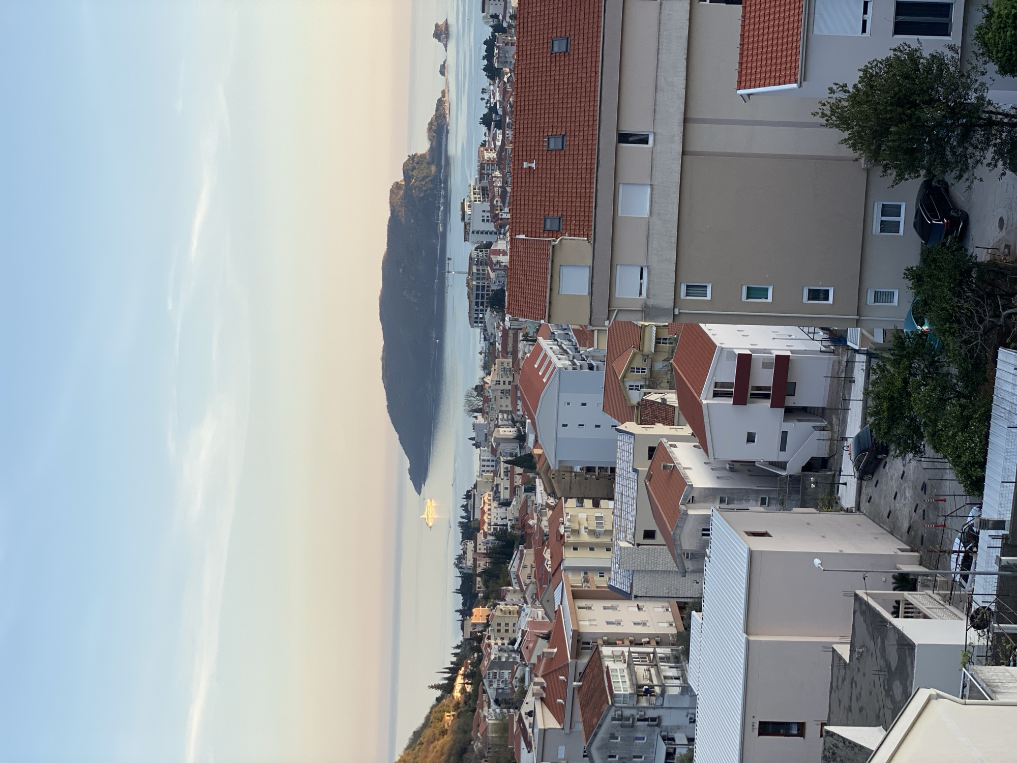 The image shows a cityscape in Budva, Montenegro, viewed from an elevated position. The scene is dominated by densely packed buildings with terracotta roofs, ranging in height from single-story structures to multi-story apartment buildings. In the background, the Adriatic Sea meets the coastline, dotted with small islands and peninsulas. A large, dark, mountainous landmass extends into the sea, likely Sveti Nikola Island. The sky is clear, with a slight gradient from blue near the top to a warm, golden hue near the horizon, suggesting either sunrise or sunset. A large cruise ship is sailing along the coast. In the foreground, rooftops, trees, and a car parked in a courtyard add depth and context to the urban landscape. The photograph appears to be taken from a high floor of a building, looking out over the city towards the sea.