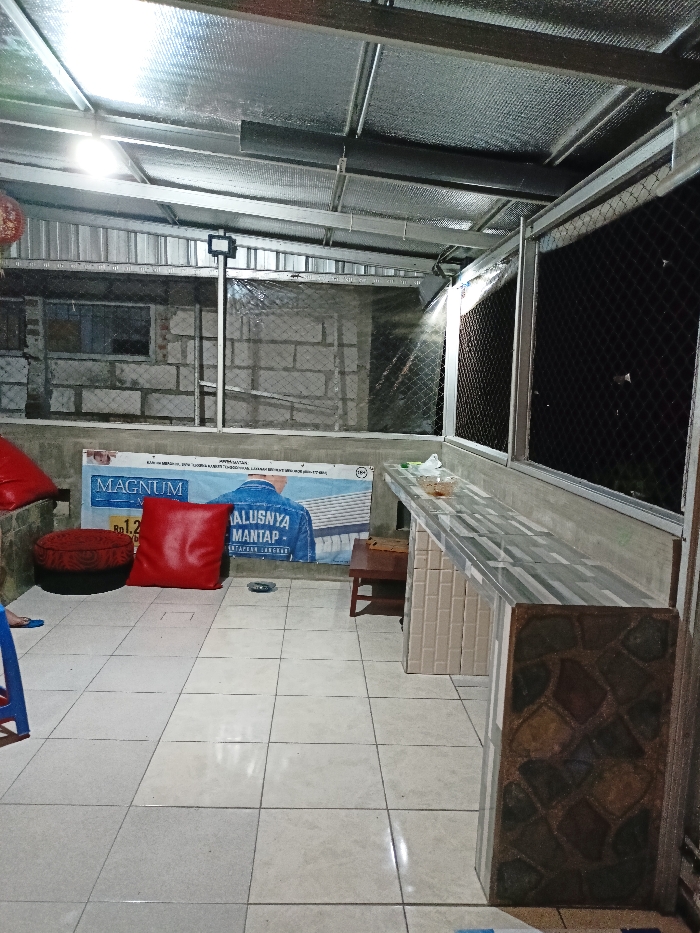 This image captures a casual, semi-enclosed space, likely a patio, rooftop area, or a small open-air establishment, illuminated by artificial lighting, indicating it is evening or nighttime. The setting appears to be in Mojosari, Indonesia.
The ceiling is constructed from corrugated metal sheets lined with reflective insulation, supported by a framework of metal beams. The floor is covered with light-colored square tiles. To the left, a casual seating area is arranged on the floor, featuring two bright red cushions – one square and one round, pouf-like – implying a relaxed atmosphere. Part of a blue plastic stool is visible nearby.
A large banner hangs on the wall behind the seating. The banner prominently features the word "MAGNUM" in white text. Other visible text in Indonesian includes "ALUSNYA MANTAP" (meaning smooth/fine and great/stable) and "SETELAH LANGKAH" (after a step), along with a price indication "Rp 1.2..." and an "18+" age restriction symbol. The wall behind the banner is constructed from unrendered concrete blocks.
To the right, a tiled counter or bar extends along the wall. Its front is adorned with a mosaic-like pattern of various-colored tiles, while the countertop features a lighter, striped tile pattern. A small wooden stool is tucked beneath the counter. A glass bowl, possibly containing snacks, rests on the counter surface. The right side of the space, beyond the counter, is enclosed by a dark wire mesh or netting, which also appears in parts of the background, acting as a barrier or screen.
No people are present, but the arrangement of seating and the counter suggests a space designed for social interaction, dining, or relaxation.