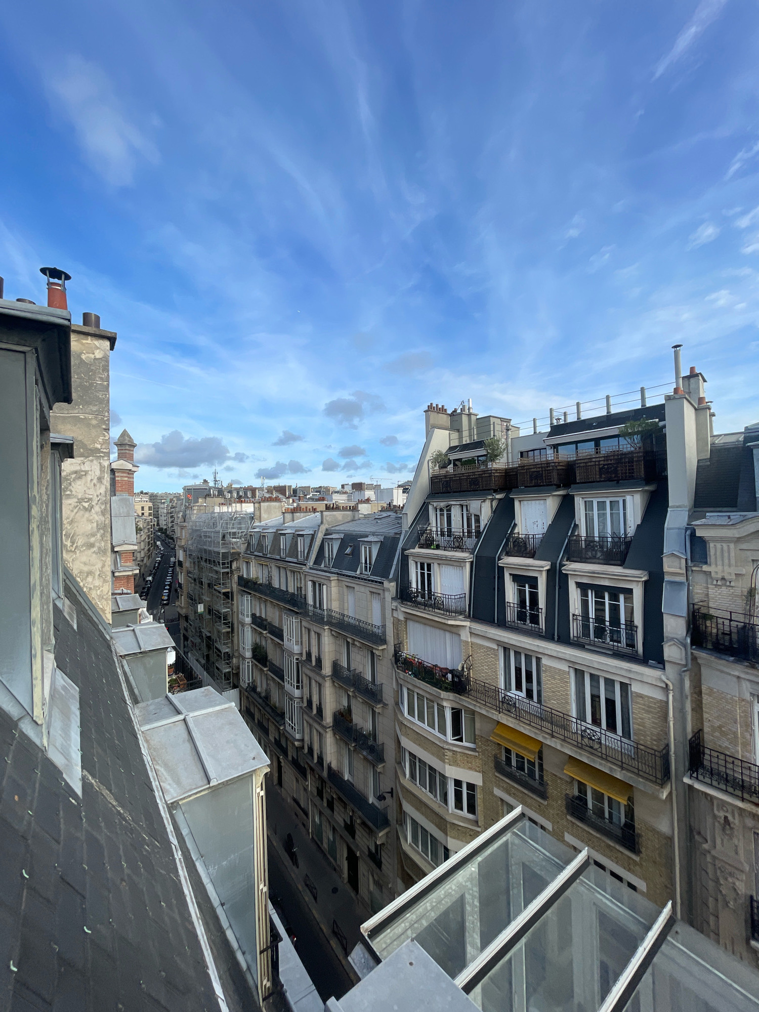 The image captures a view of Parisian rooftops and a street below on a clear day. The perspective is from a high vantage point, looking down and across a row of classic Haussmannian buildings. The architecture is characterized by mansard roofs with dormer windows, intricate iron balconies, and stone facades. Chimneys are visible on many of the rooftops, as are various rooftop terraces with greenery and railings.
The street below is lined with parked cars, indicating an urban setting. The buildings on either side of the street are of similar architectural style, creating a sense of uniformity typical of Paris. The sky is bright blue with scattered cirrus clouds. The overall scene is one of a vibrant, lived-in city, with a clear and crisp atmosphere. The presence of scaffolding on one building suggests ongoing renovation or construction. The image appears to be taken in a residential district of Paris.