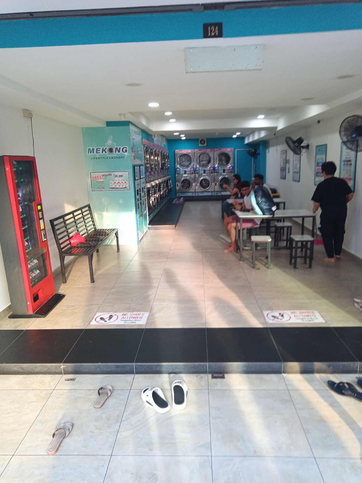 The image depicts the interior of a laundry shop with a vending machine on the left and rows of washing machines and dryers in the background. A bench is placed in front of the washing machines, with a red bag on it. Several people are present, some sitting at tables and others standing. A sign on the floor indicates "NO SHOES ALLOWED." The environment appears to be clean and well-lit, with tiled flooring and bright overhead lighting. The shop is located in Kuala Lumpur, Malaysia, as suggested by the signage and the presence of a vending machine common in the region.