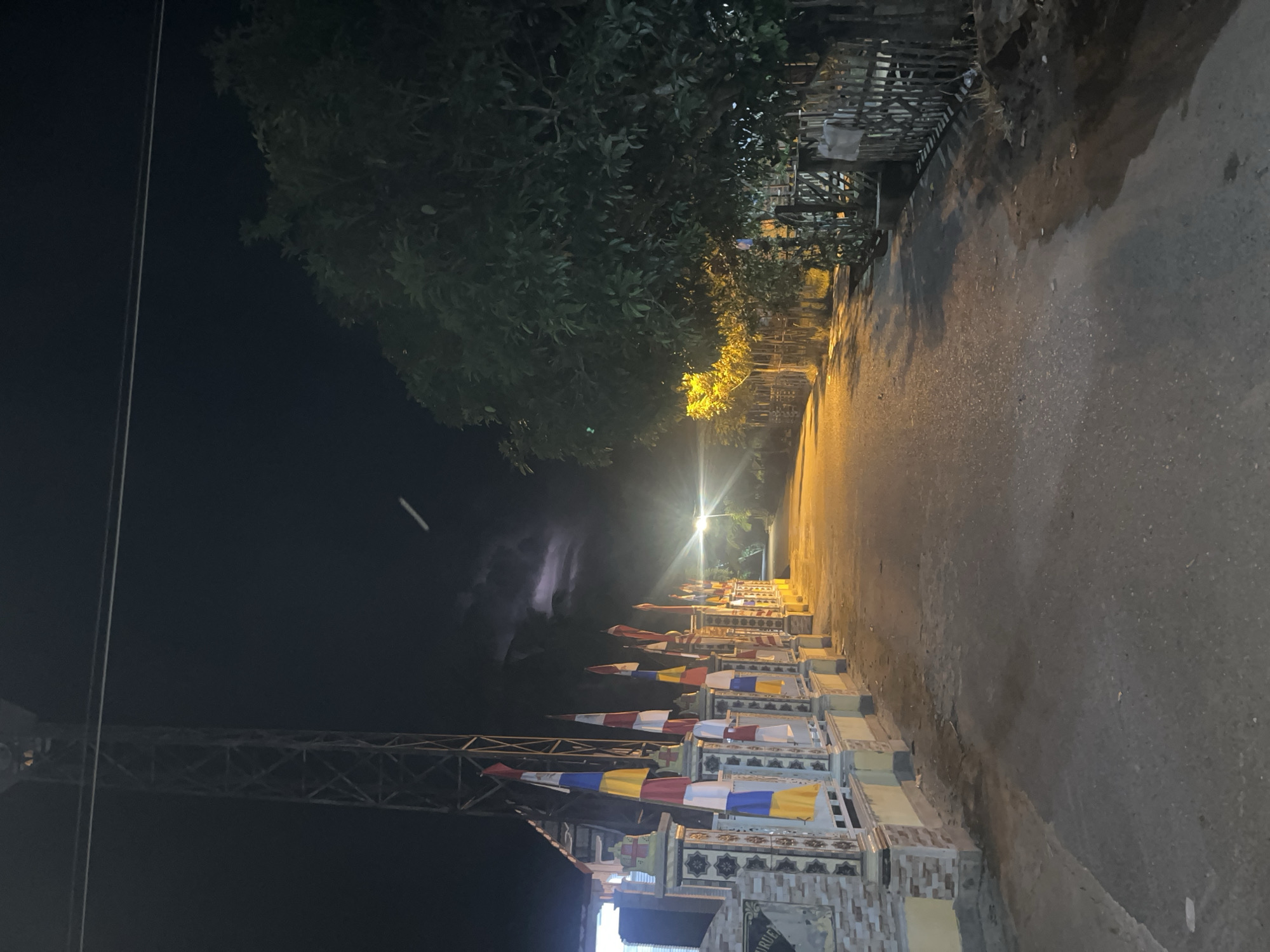 A nighttime view of a street in Blangseureukuy, Indonesia shows a road illuminated by a bright street light. The road is paved and appears wet in places, reflecting the light. On the left side of the road, there is a decorative white fence with colorful flags and banners hanging from it. Behind the fence, a building is visible, along with a tall metal tower. A large tree with dark green foliage stands on the right side of the road, with a wooden fence running along its base. The sky is dark with some faint cloud cover, and a bright, possibly lunar, light source is visible in the upper portion of the frame. A thin, white elongated object is visible in the sky, possibly an airplane or a streak of light.