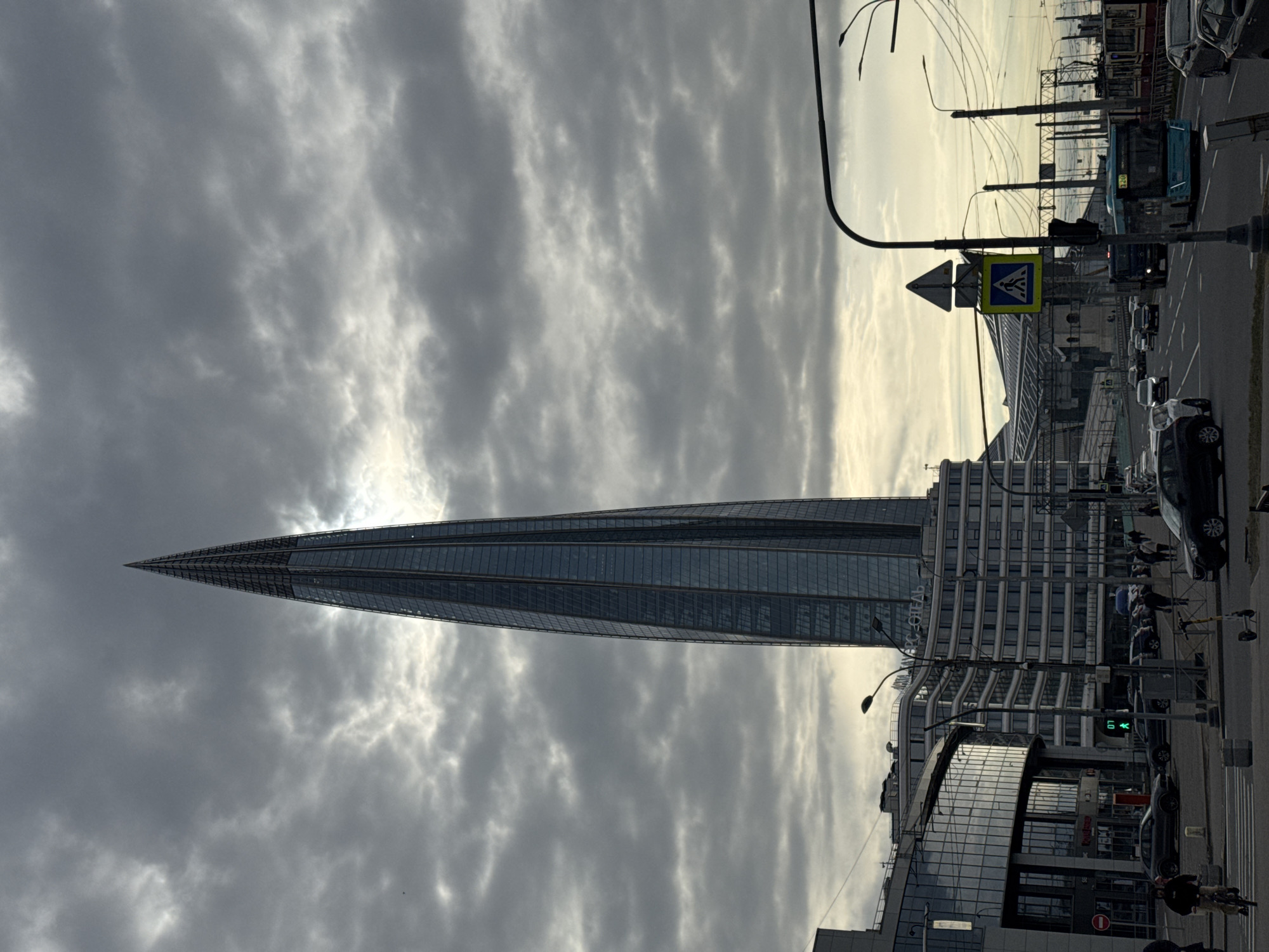 The image shows the Lakhta Center skyscraper in Saint Petersburg, Russia, under a cloudy sky. The skyscraper has a needle-like design, tapering to a sharp point. The surrounding buildings and street scene indicate a urban environment, with cars, pedestrians, and a crossing sign visible along a street. The presence of overhead cables and street lights further suggests an active urban street.