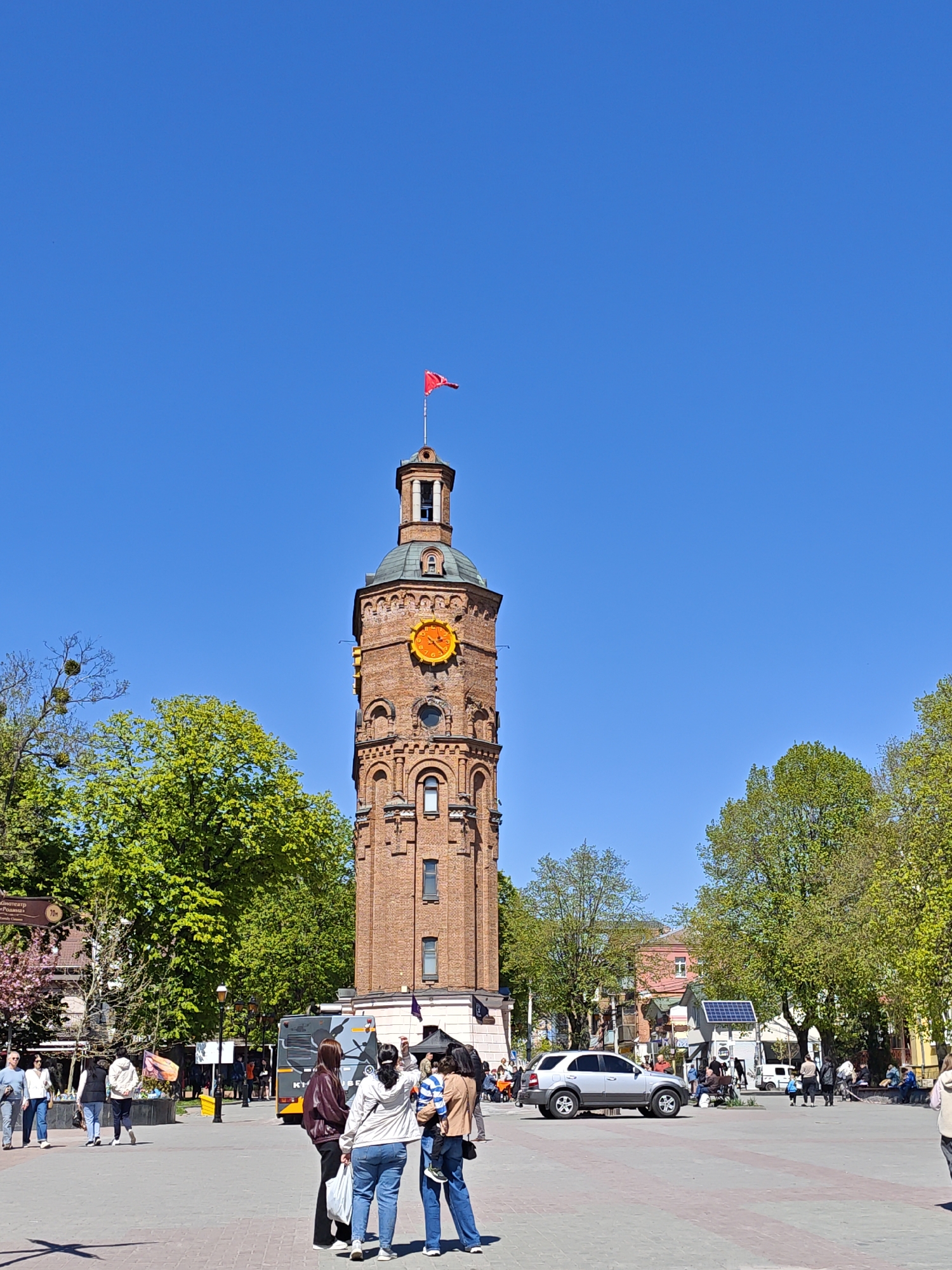 The image captures a sunny day in the city of Vinnytsia, Ukraine, focused on the iconic Water Tower. The brick tower, topped with a green dome and a red flag, stands prominently in the city center. A large clock is visible on the tower's façade.
In the foreground, several people are gathered on the paved square. One group is taking photographs of the Water Tower, with one person holding a small child. Other pedestrians are walking through the square in various directions.
Behind the group, a grey bus with Cyrillic lettering is visible. A silver SUV is parked on the square. Trees with lush green foliage frame the scene. In the background, other buildings and street furniture are present, including a small structure with a solar panel. The sky is a clear, bright blue.