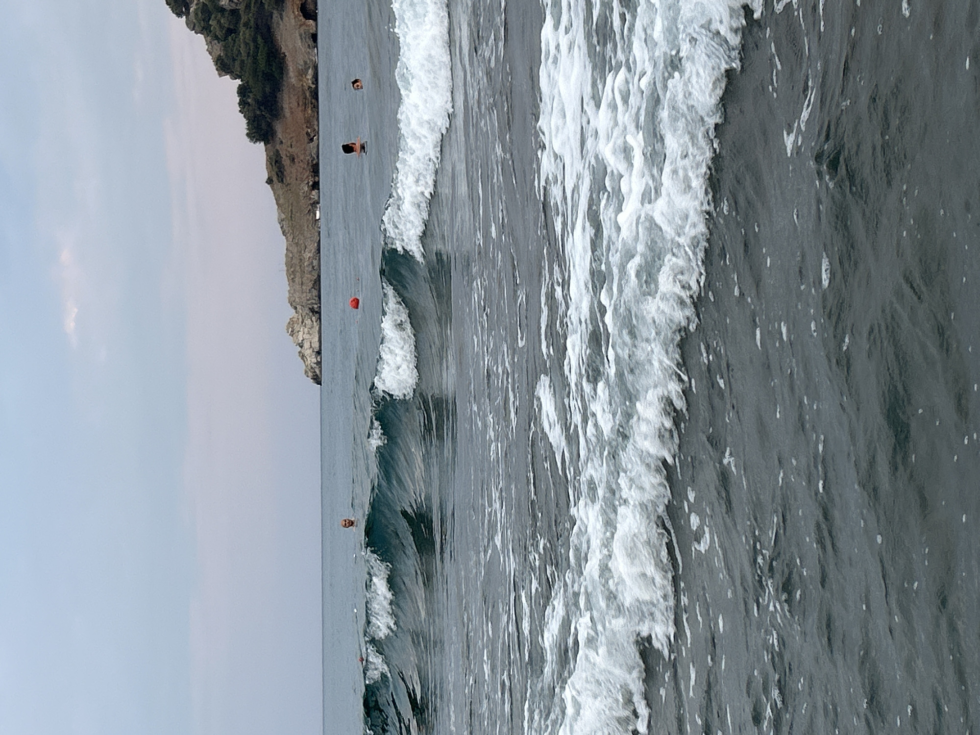 This is a medium-shot, eye-level, horizontal, outdoor, daytime photograph taken at Iztuzu Beach in Ortaca, Turkey. The image captures a section of the sea with several waves rolling towards the shore. In the mid-ground, a person's head is visible above the water, seemingly swimming. Further to the right, two other individuals are partially submerged, also appearing to be in the water. A red buoy floats in the water to the left of the frame. In the background, a rocky, tree-covered hill slopes down towards the sea. The sky is overcast with a pale blue hue, and the sea is a dark, choppy gray-blue. The focus is on the water and the people within it, with the shoreline and vegetation forming a backdrop.