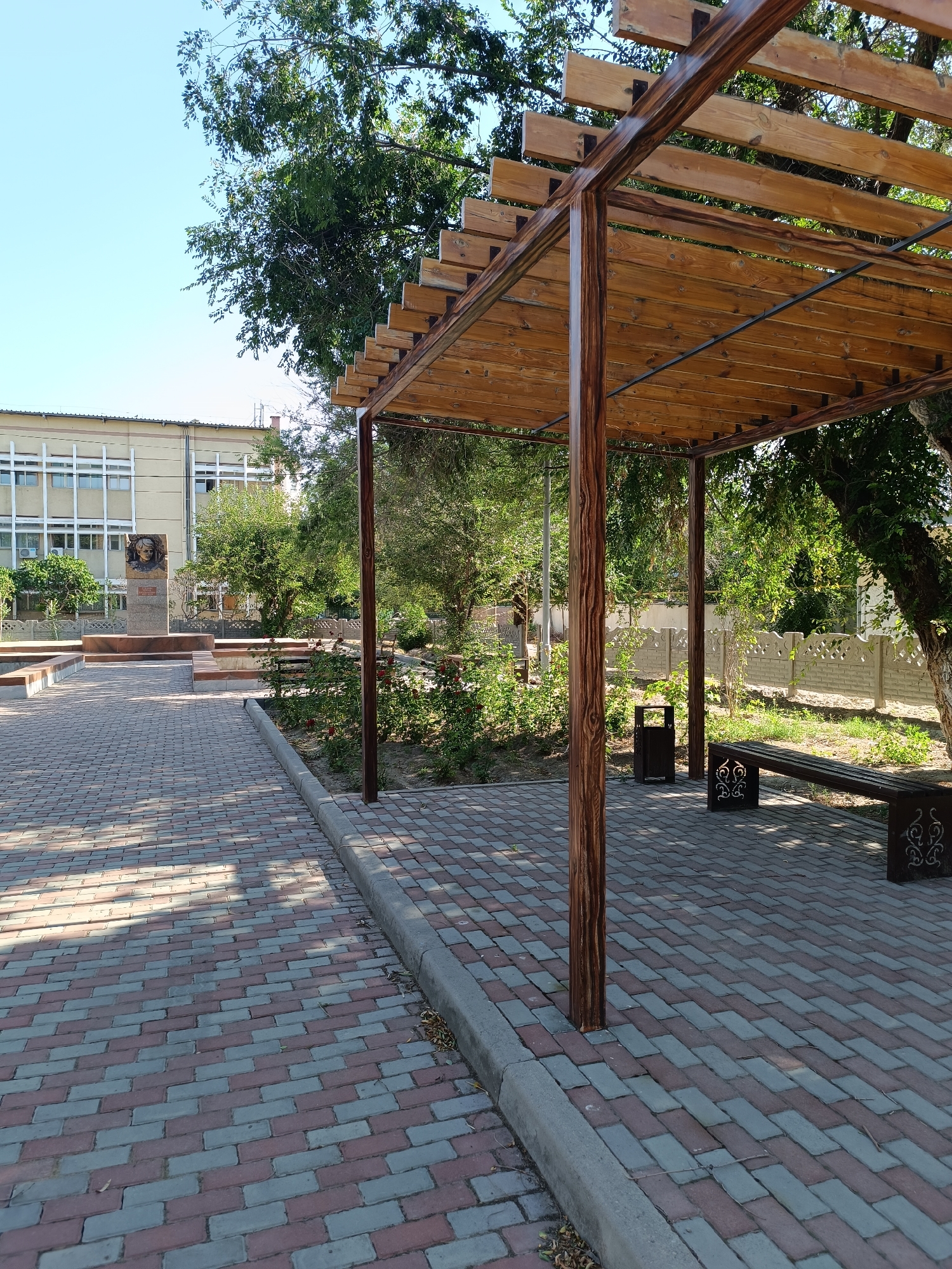 The image depicts an outdoor public space with a paved walkway made of red and gray bricks. To the right, a wooden pergola with a slatted roof provides shade over a portion of the walkway. Beneath the pergola, a dark-colored bench with decorative cutouts and a trash receptacle are visible. A concrete curb separates the paved area from a grassy area with plants and trees.
In the background, a light-colored building with multiple windows is visible, suggesting it might be an administrative or educational institution. To the left of the building, a monument or memorial with a bust of a person is situated on a pedestal. A low, decorative fence runs along the perimeter of the property. The overall scene appears to be a well-maintained public area, likely within a city, given the presence of the building and monument. The environment is bright and sunny, indicating daytime. There are no people visible in the image.