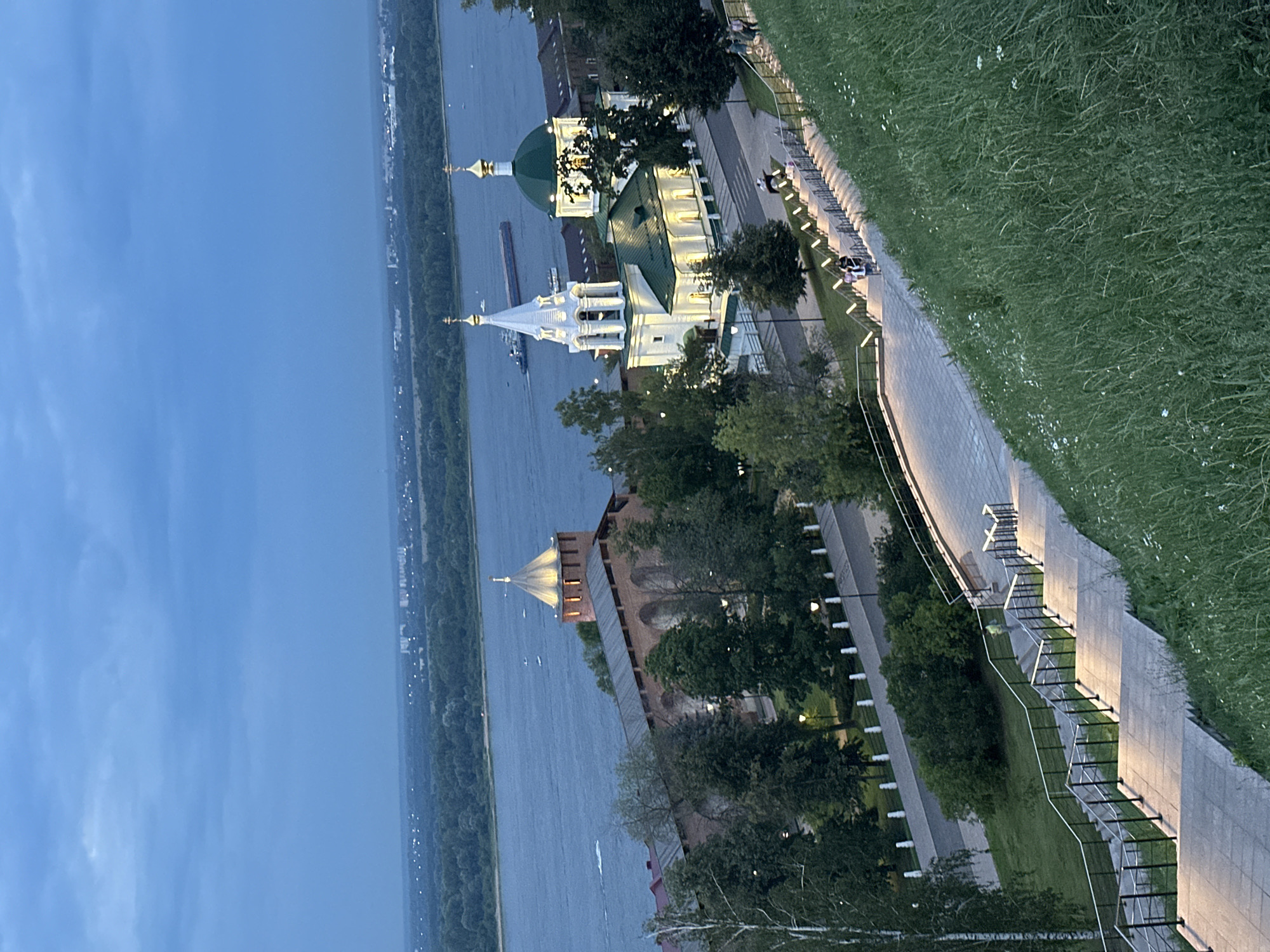 The image shows a panoramic view of Nizhny Novgorod, Russia, at dusk. In the foreground, a grassy slope leads down to a wide, illuminated stone staircase with black railings. Several people are walking up the stairs. Further down, a tree-lined walkway borders the Volga River.
On the riverbank, a historic brick fortress wall with an arched design is visible, topped by a conical tower illuminated from within. Adjacent to the fortress is a brightly lit white church with green domes and golden crosses, featuring a tall bell tower. A barge is sailing on the wide, dark blue river. In the distance, a city skyline with trees forms the horizon under a dusky blue sky with scattered clouds. The overall scene suggests a picturesque evening at a historical landmark.