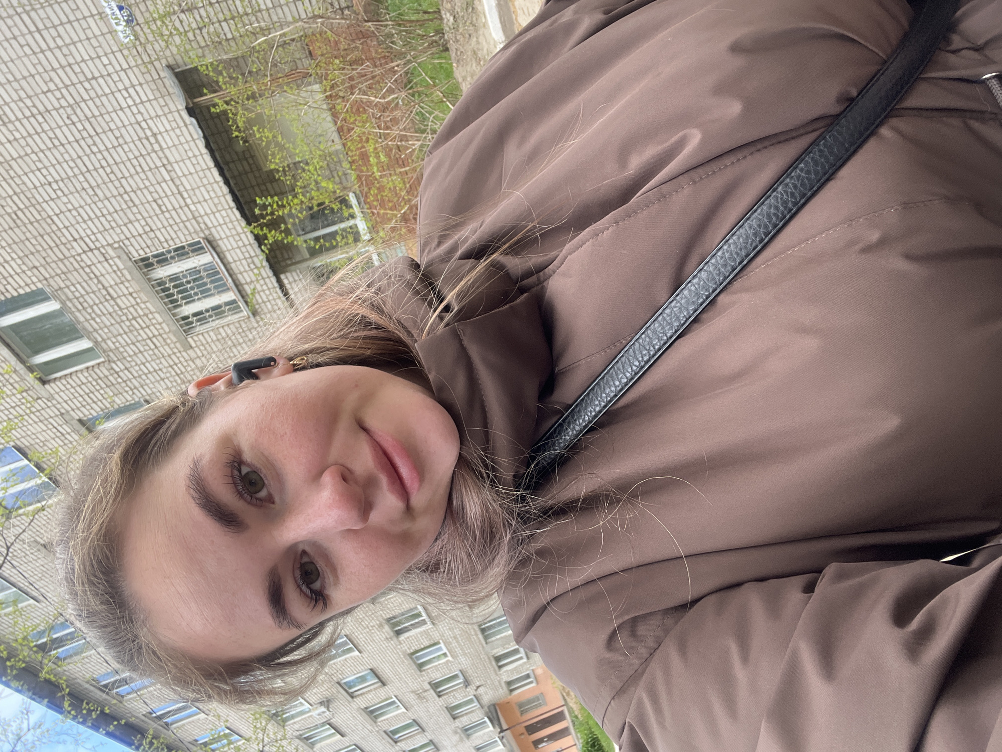 A young woman with fair skin, light brown hair, and defined eyebrows takes a selfie outdoors in Chaikovsky, Russia. She is wearing a brown puffy jacket and a black purse strap is visible across her body. Behind her is a multi-story apartment building made of beige brick, with rectangular windows and sprouting green foliage. The photograph appears to be taken during the day in early spring.