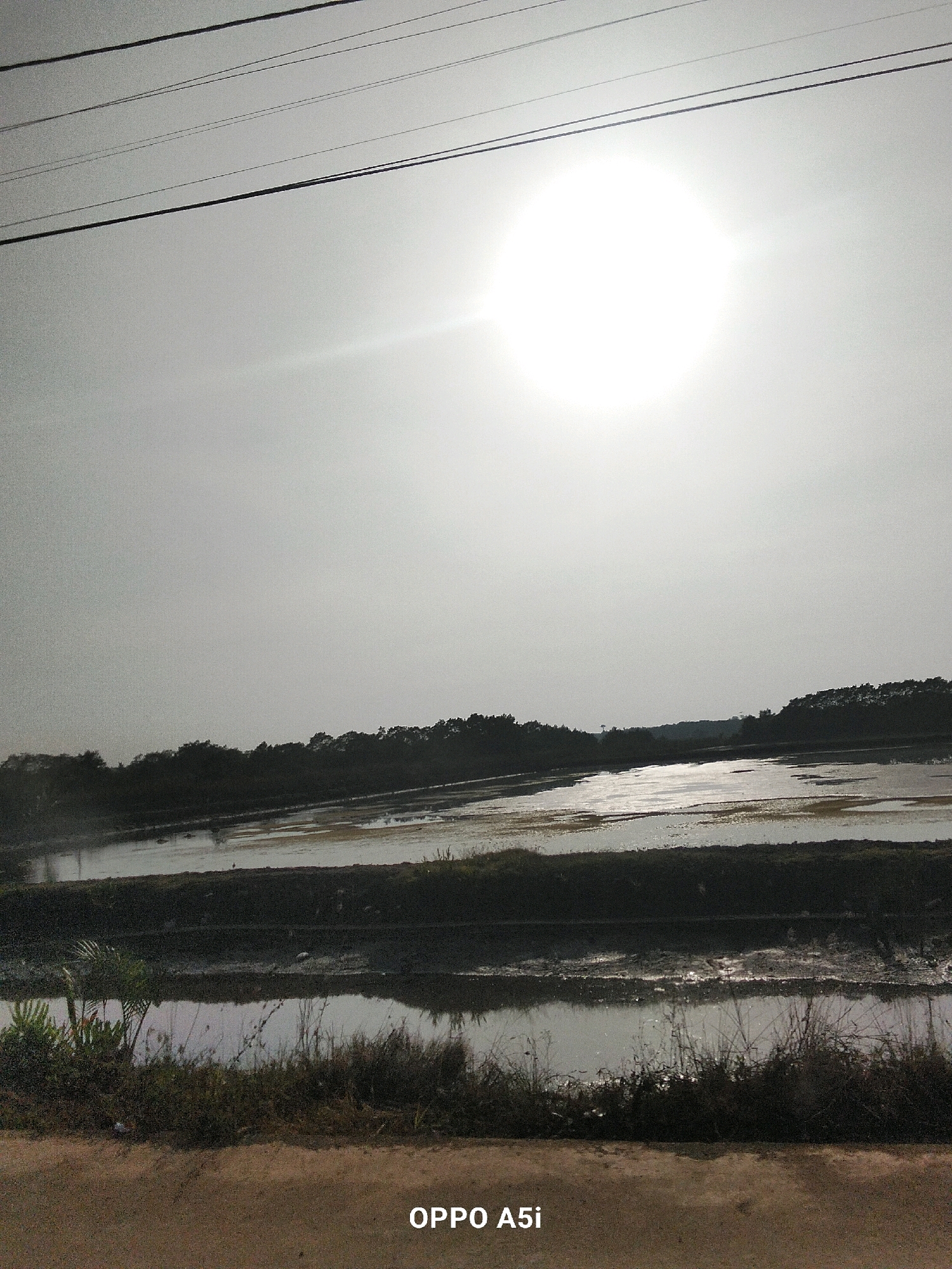 The image depicts a landscape scene with a bright sun in a hazy sky. Power lines are visible stretching across the top of the frame. Below the sky is a dark silhouette of trees and foliage, with bodies of water reflecting the sky in the foreground. The water appears to be in flooded fields, possibly rice paddies, with muddy banks and some sparse vegetation along the edges. The ground in the immediate foreground is a dusty, light brown surface. The text "OPPO A5i" is watermarked in the bottom right corner. The scene is likely outdoors in a rural or semi-rural area. While the image is identified as being from Tondowatu, Indonesia, no specific landmarks or elements within the image confirm this location.