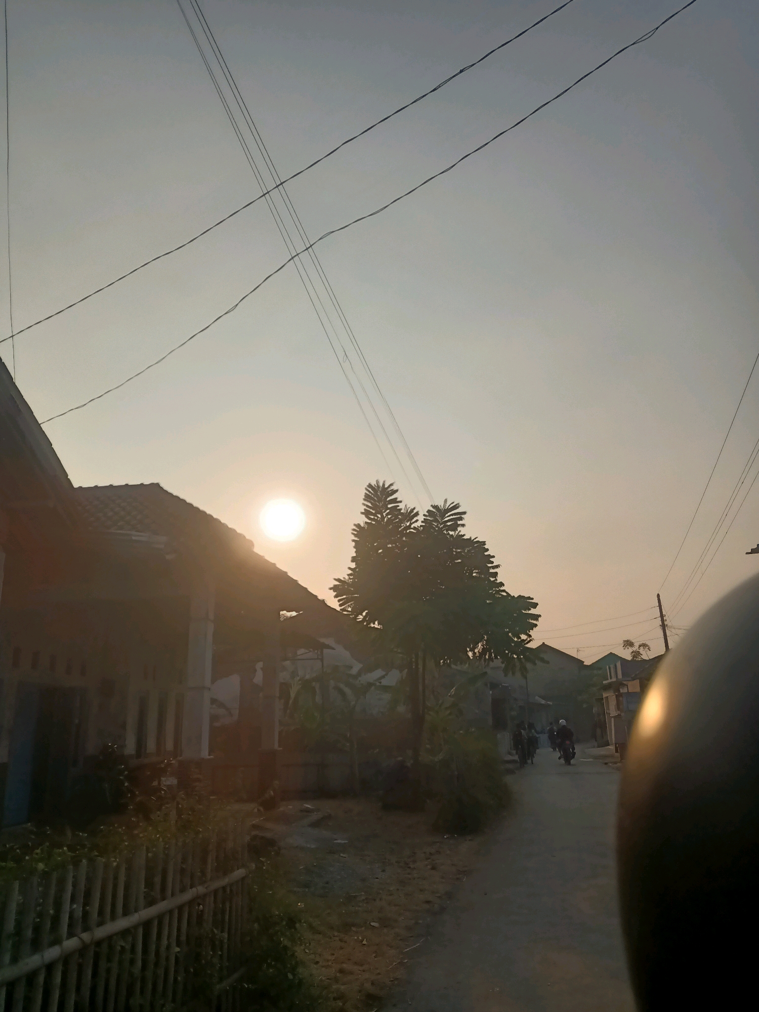 This image captures a street scene in Pecabean, Indonesia, during what appears to be sunset or sunrise. The sky is a hazy, pale orange, with several thin electrical wires crisscrossing overhead. On the left side of the frame, a traditional Indonesian house with a dark tiled roof and a covered porch is visible, partially silhouetted against the bright sun. The house is bordered by a bamboo fence and overgrown greenery. A dirt road runs through the center of the image, with several people on motorcycles riding away from the viewer. The road is lined with houses and trees. In the foreground on the right, a portion of a dark, rounded object, possibly a helmet, is in focus, suggesting the photo was taken from the perspective of someone riding a motorcycle. The overall atmosphere is rural and peaceful.
