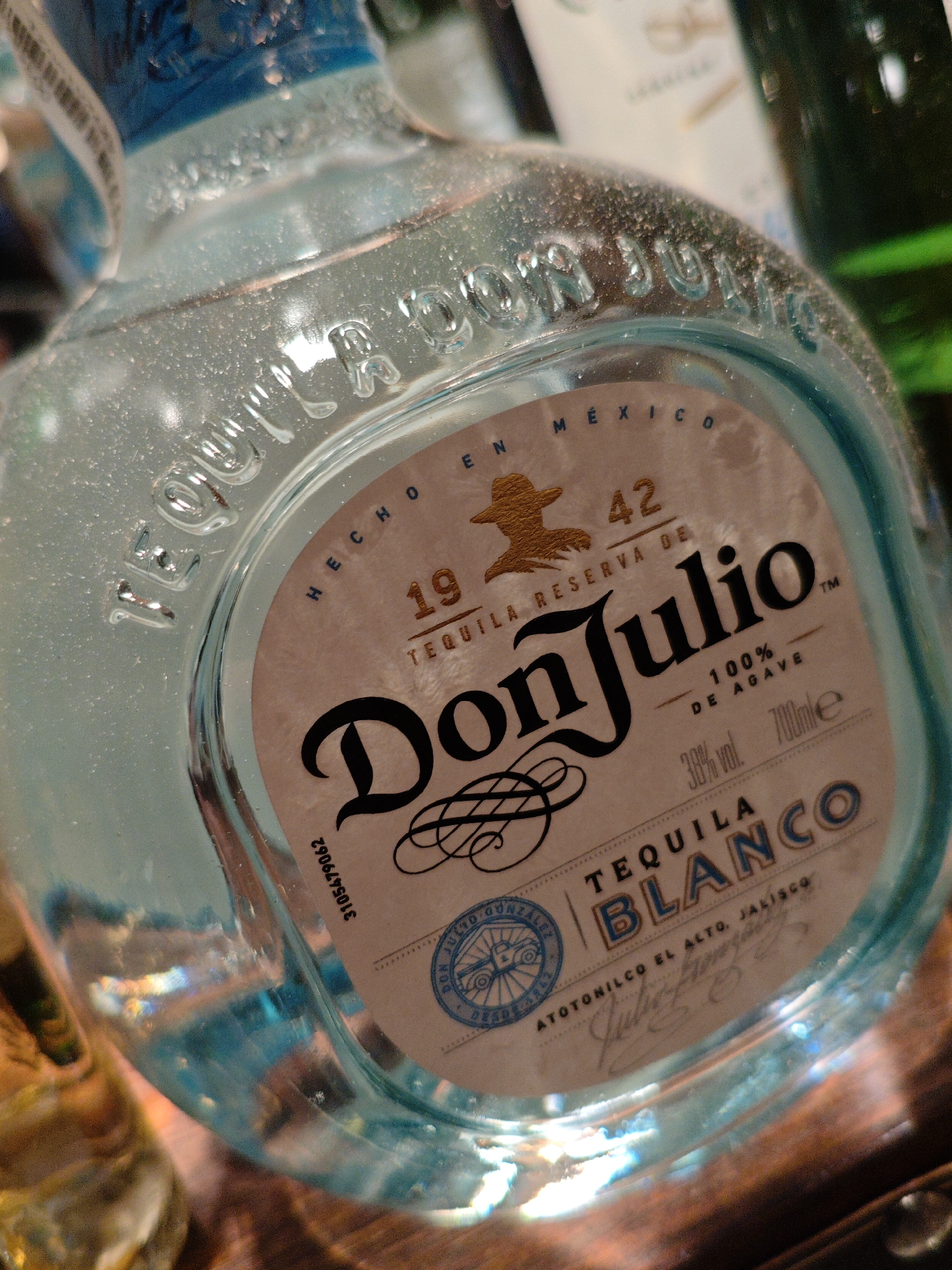 A close-up, low-angle shot captures a clear glass bottle of Don Julio Tequila Blanco, positioned in the foreground. The bottle is partially filled with a pale blue liquid, and the lighting creates highlights and reflections on its surface. The label is prominent, featuring the brand name "Don Julio" in large, stylized black lettering, with "TEQUILA BLANCO" below it. Several details are visible on the label, including "100% DE AGAVE," "38% vol," and "700ml." The label also contains a seal with a sun-like symbol and text, and a smaller code reading "3105479062." The bottle's glass is embossed with lettering that partially reads "DON JULIO." In the background, out of focus, are other bottles and possibly shelves, suggesting an indoor setting such as a bar or liquor store. The image is devoid of people or any contextual information about its location in Kyiv, Ukraine.