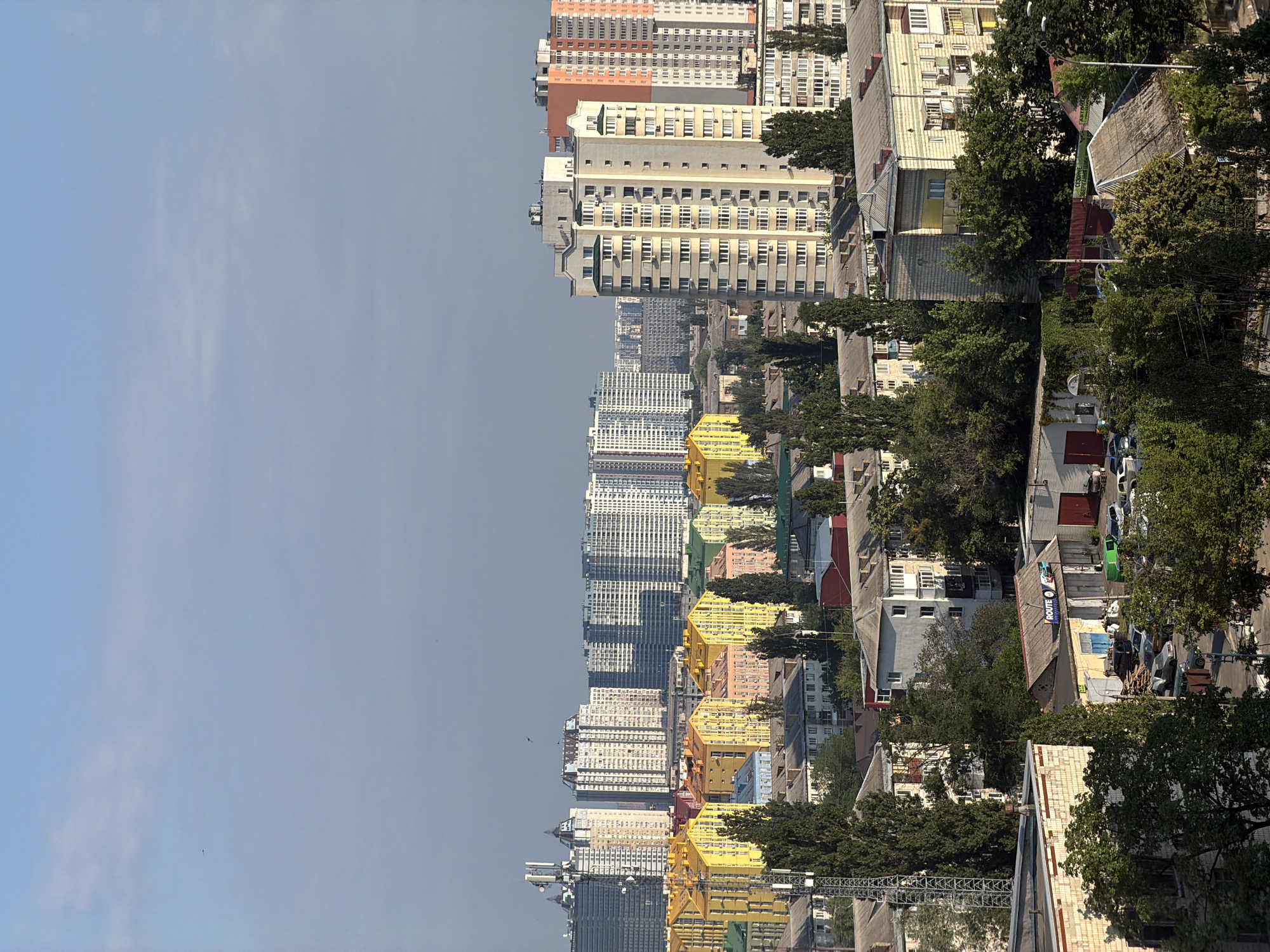 This is a high-angle, full shot, outdoor, daytime photograph of a cityscape in Kyiv, Ukraine. The image features a wide expanse of the sky dominating the upper portion, with a few wispy clouds present. Below the sky, a dense collection of buildings fills the frame. In the background, several tall, modern apartment buildings are visible, some in white with many windows, others in grey or with colorful accents. In the middle ground, a striking row of brightly colored apartment buildings stands out; these buildings are painted in vibrant shades of yellow, orange, pink, and green, creating a distinctive and cheerful architectural style. Interspersed between and in front of these structures are numerous trees, varying in height and density. Closer to the foreground, more modest, older residential buildings and some industrial or commercial structures are visible, with some featuring grey, tiled roofs. Several cars and what appear to be utility poles are scattered throughout the scene. The overall impression is one of urban development with a mix of architectural styles and significant green spaces provided by trees.