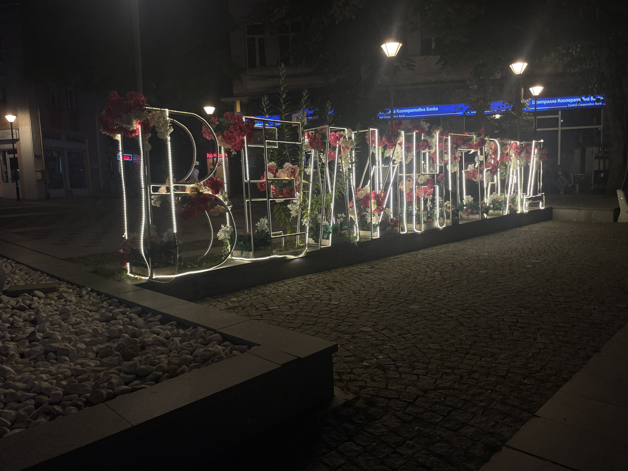 A nighttime, eye-level exterior shot shows a large, illuminated art installation in Velingrad, Bulgaria. The installation spells out the word "BEAUTY" in large, block letters, each outlined in white LED lights and adorned with a variety of artificial flowers, primarily in shades of pink and white, with some greenery. The letters are constructed from a metallic frame. In front of the installation, a raised planter bed is filled with white, smooth pebbles and a few small green plants. The ground is paved with dark cobblestones, which are wet and reflect the ambient light. In the background, buildings with illuminated windows are visible, along with streetlights casting a warm glow. A blue banner for "Central Cooperative Bank" is mounted on one of the buildings. The overall scene is urban and appears to be in a public square or pedestrian area.