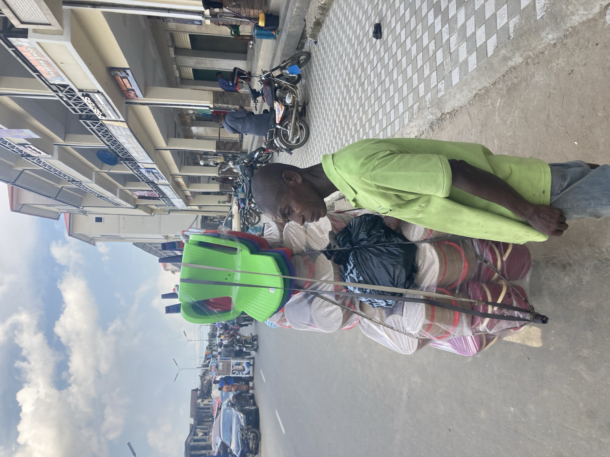 This image captures a bustling street scene in Umuode, Nigeria, during the daytime under an overcast sky. In the foreground, a dark-skinned man, appearing middle-aged, wears a light green long-sleeved shirt and blue jeans. He is focused on the goods he is transporting, looking downwards.
The man is pushing or pulling a cart heavily laden with various items. Prominently stacked on the cart are several bright green plastic chairs, along with others in red and purple, all wrapped in clear plastic. Below the chairs, numerous circular, flat items are visible, individually wrapped in plastic. These items appear to be mats or possibly hats, coming in shades of beige, maroon, and purple. A black plastic bag is also secured to the cart.
The background reveals a lively urban environment. To the left, a street stretches into the distance, populated by motorcycles and cars, indicating active traffic. Alongside the street are multi-story buildings housing various shops. Shop signs are visible, with readable text including "FASHION SHOP," "SISTERS," and "BEST OF FASHION." The sidewalk in front of these shops features a distinctive checkered pattern of light and dark tiles. Other people are visible further down the street, some on motorcycles, contributing to the busy atmosphere of a commercial district. The overall scene suggests a typical day of commerce and movement.
