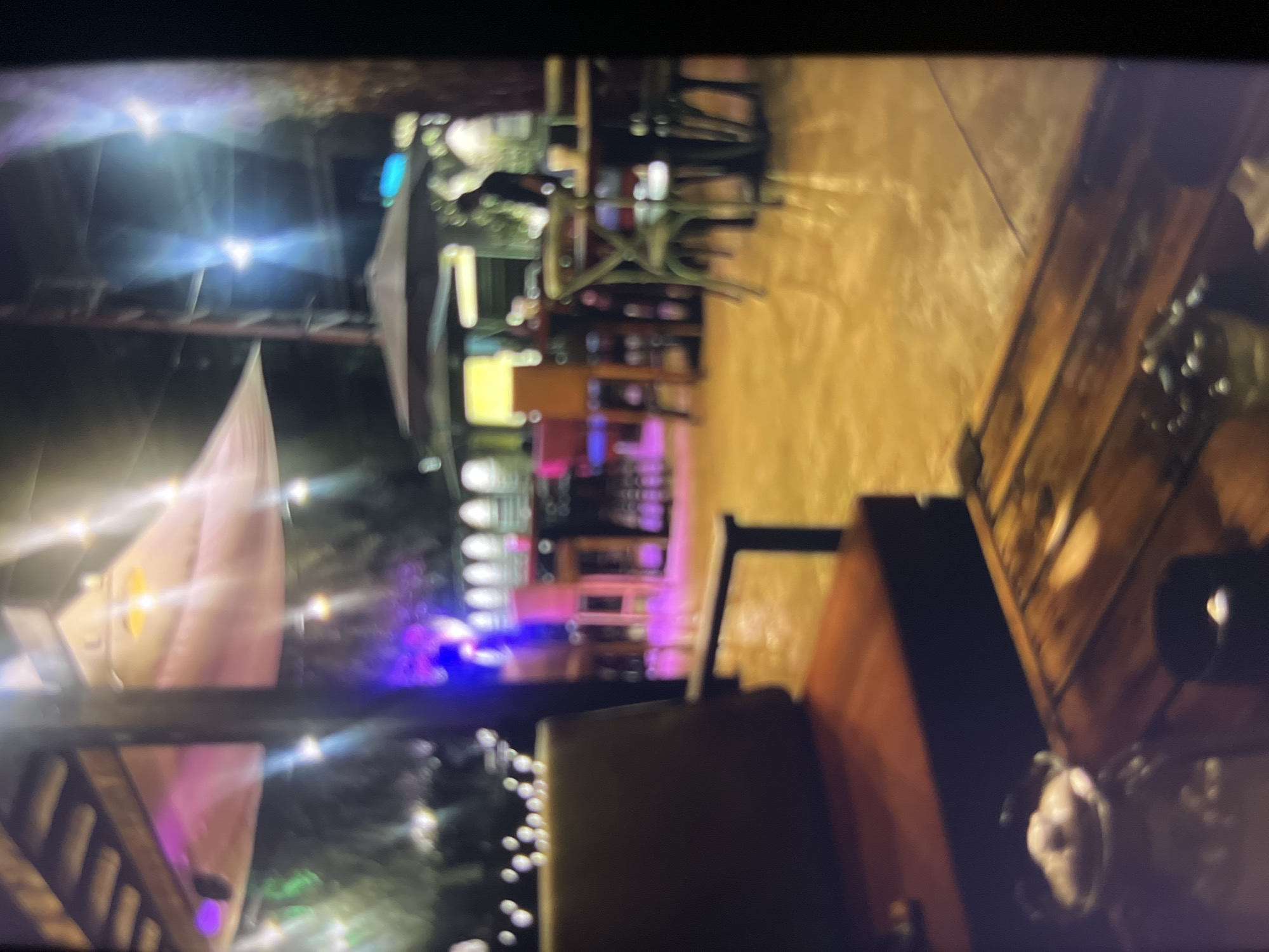 The image depicts an outdoor seating area of a restaurant or bar at night. The scene is illuminated by string lights and overhead lamps, casting a warm glow on the surroundings. In the foreground, a wooden table is visible with some objects on it, including what appears to be a candle in a holder. To the left, a plush chair with a dark cushion and a wooden frame is positioned. The background reveals more seating arrangements, including tables and chairs, as well as umbrellas for shade. There are also some figures blurred in the distance, suggesting patrons or staff. The overall ambiance is casual and inviting, hinting at a lively establishment. The location is identified as a city in Nigeria, specifically Udu.