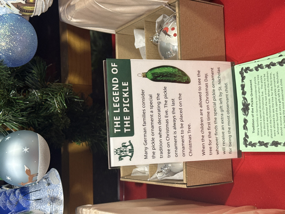 The image shows a Christmas ornament set featuring "The Legend of the Pickle". The display includes a card explaining the tradition: German families place a pickle ornament on the Christmas tree last. On Christmas Day, the child who finds the pickle first receives an extra gift from St. Nicholas for being observant. The set also contains a pickle-shaped ornament. A second card elaborates on the tradition of the pickle ornament.