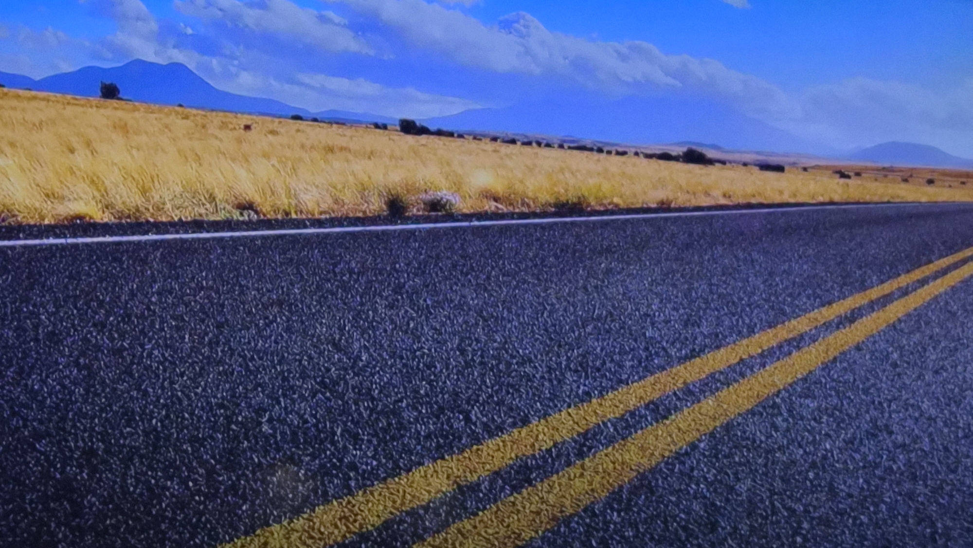 The image depicts a rural asphalt road with two yellow lines marking the lane. The road runs through a landscape of dry, yellow grassland. In the background, there are rolling hills and mountains under a blue sky with scattered clouds. The location is not Kalipetrovo, Bulgaria.