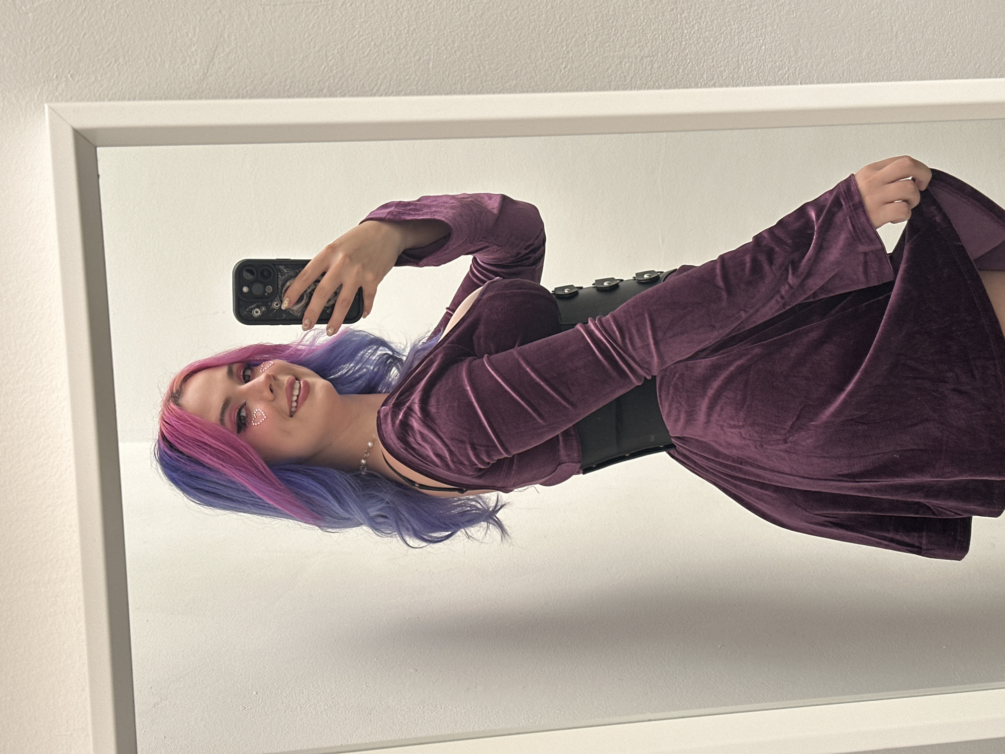 A woman is posing for a mirror selfie. She is wearing a long-sleeved, purple velvet dress and a wide black corset. Her hair is dyed with purple and blue streaks, and she has heart-shaped glitter stickers on her face. She is holding a black smartphone in her right hand, and the phone case has a detailed design. The mirror has a white frame. The wall behind the mirror is white. The location is not discernible from the image.