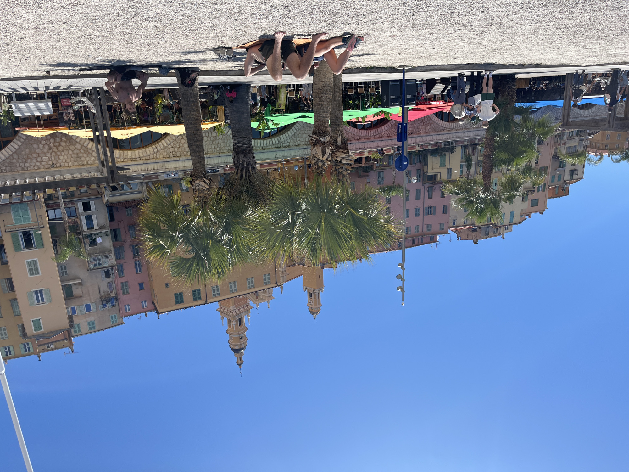The image is taken from the perspective of someone on the ground, looking up at an angle. The bottom portion of the image is blue sky. The top portion shows the buildings and other structures in Menton, France, including the prominent Baroque Basilica of Saint Michael Archangel with its bell tower. In the foreground, near the apparent horizon, are palm trees and a number of people. Some individuals are standing or walking, while others are lying on the ground sunbathing. Beach pebbles are visible on the ground in the foreground. Behind the people and palm trees are shops or restaurants with canopies in a variety of colors, adding to the vibrant scene.