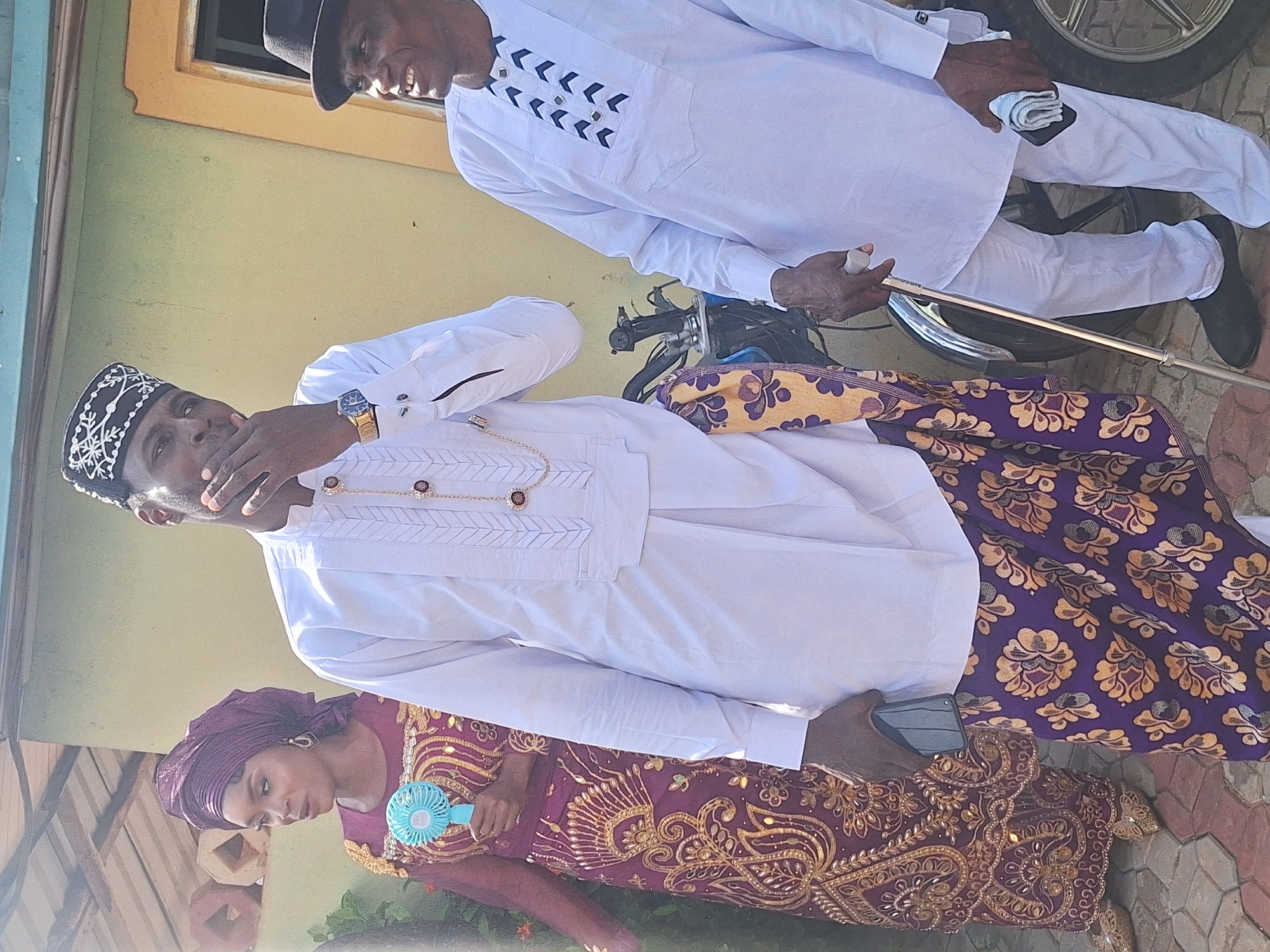 The image depicts three individuals in Rido, Nigeria. An older man wearing a white traditional Igbo attire with black embellishments, a black fedora hat, and black shoes is partially visible at the top. He is holding a walking stick and what appears to be a handkerchief or small cloth. Below him, a man in a white Igbo-style shirt with brown and gold embellishments and a dark-colored cap with white embroidery is seen covering his mouth with his hand. He wears a gold chain and a watch. He also wears a dark-patterned fabric wrapped around his waist. At the bottom of the image, a woman in a purple headscarf and a purple and gold embroidered dress is holding a small blue fan. The background shows a light yellow wall. They stand on a tiled pavement. The image seems to have been taken during a sunny day.