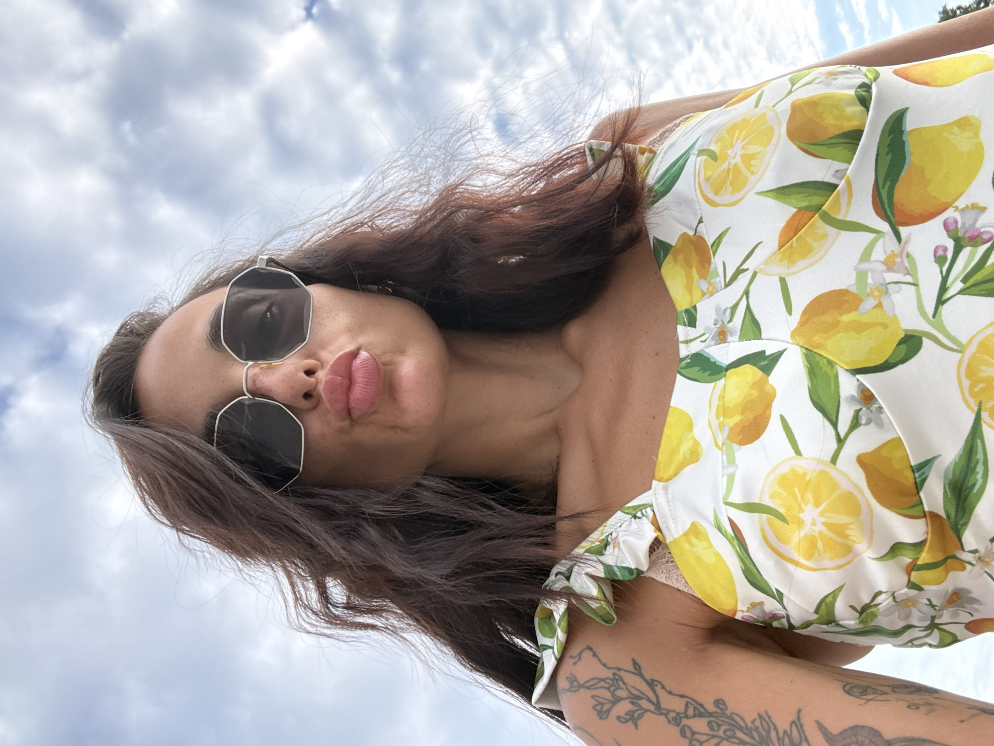 A low-angle, close-up outdoor selfie shows a woman with dark, wavy hair wearing sunglasses and a white top with a lemon pattern. She is puckering her lips. A tattoo of a floral design is visible on her left arm. The background consists of a cloudy sky. The image does not provide any information about the city of Pleven, Bulgaria.