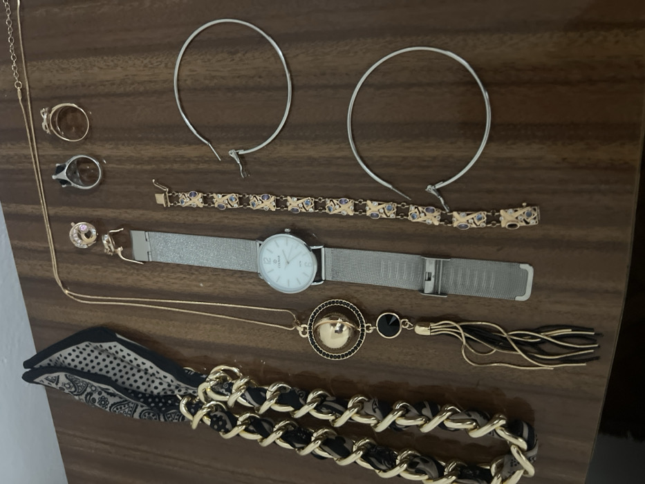 Multiple items of jewelry and accessories are arranged on a polished, medium-brown wooden surface, exhibiting a vertical wood grain pattern. The scene is located in the city of Smederevska Palanka Municipality, Serbia.
At the top left, a folded scarf, predominantly black and off-white with a patterned design, is positioned, attached to a thick, gold-colored chain interwoven with dark fabric. Below this, a long, gold-colored necklace features a circular pendant with a central gold sphere, a ring of small black beads, and a hanging black stone from which gold and black tassels extend.
Centrally placed is a silver-colored wristwatch with a white face displaying "COLMAR" and an unclasped silver mesh strap. To its right, an unclasped gold-colored bracelet features alternating rectangular and oval links, with the rectangular links inlaid with small blue or purple stones.
At the top right, a thin, gold-colored chain necklace is laid horizontally. Below it, from left to right, are a small gold-colored stud earring with a pink stone, a silver-colored ring with a dark stone set in a crown-like bezel, and a plain gold-colored band ring.
Two large, unfastened silver-colored hoop earrings are positioned vertically stacked on the bottom right of the frame.