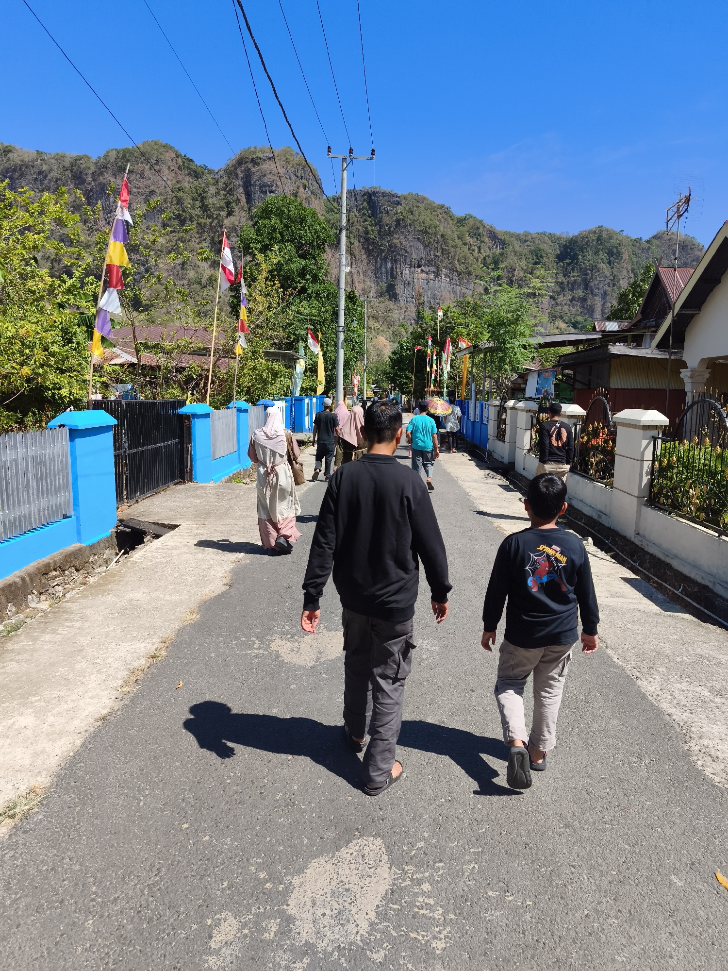 A group of people are walking down a paved road under a clear blue sky in Tonasa, Indonesia. The road is lined with a blue fence on the left and houses with ornate fences on the right. Several flags are displayed along the road, and lush green trees and rocky hills form the background. Two young boys in black t-shirts are walking in the foreground, facing away from the camera. The overall scene depicts a sunny day in a village setting.