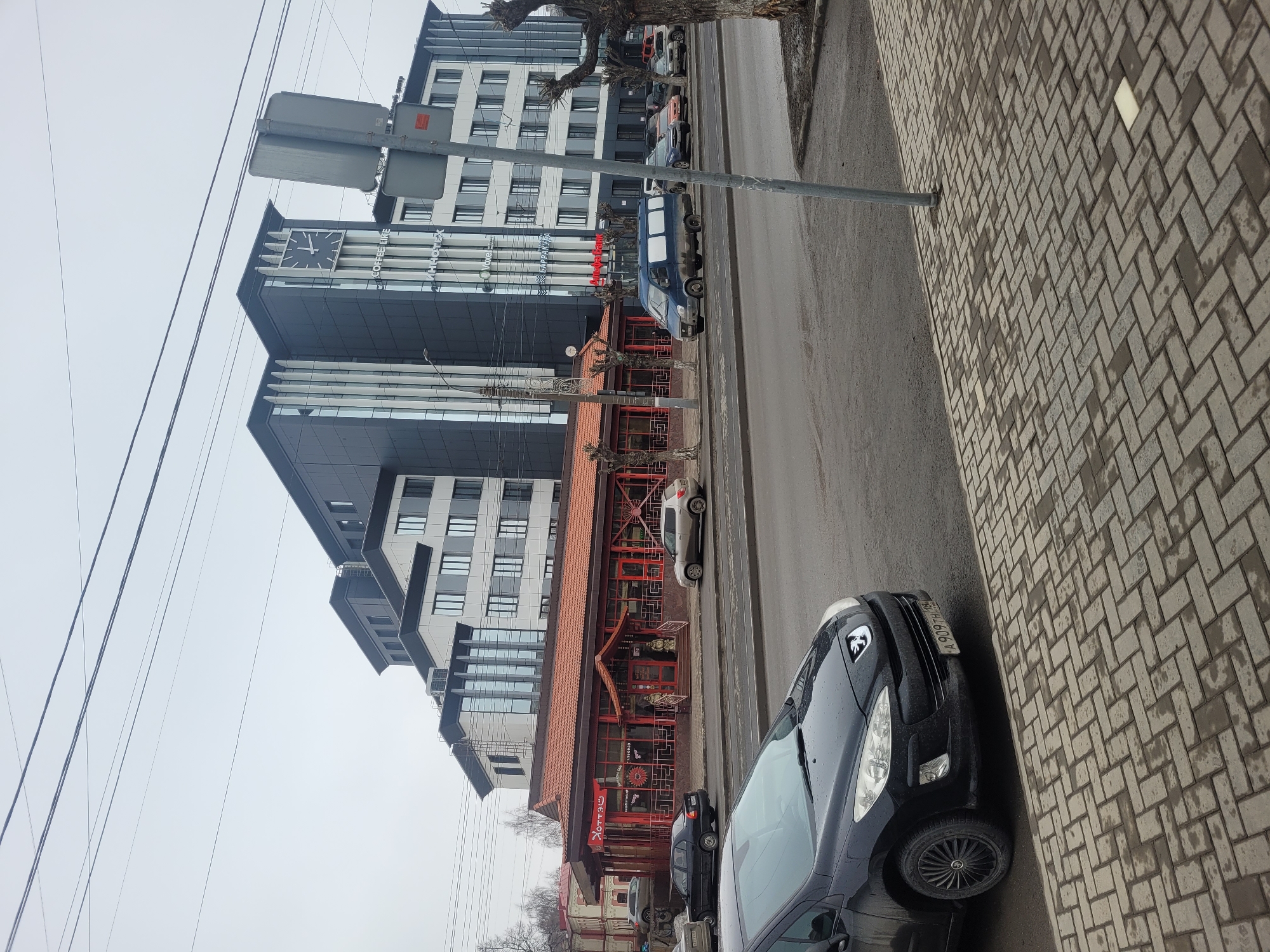 The image depicts a street scene in Izhevsk, Russia, characterized by a mix of modern and traditional architectural elements. In the background, a modern building with a clock tower is visible, contrasting with a traditional-style restaurant featuring a red tile roof and Asian-inspired decor. Several cars are parked along the street, including a black Peugeot with license plate FH 606 Y. The foreground shows a brick-paved sidewalk, typical of urban areas. Power lines are strung overhead, contributing to the urban landscape. The weather appears overcast, suggesting a cloudy day. The image captures a blend of contemporary and cultural features within the cityscape of Izhevsk.