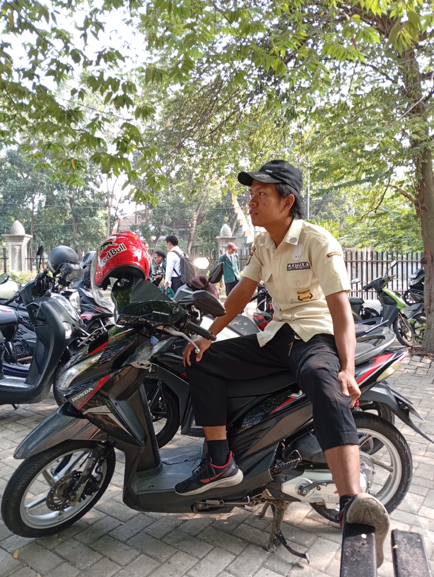 A man wearing a beige uniform and a black baseball cap sits on a black and red scooter parked on a paved area. He is positioned on the right side of the scooter, facing left. The uniform shirt has patches and text. His left hand rests on the scooter's handlebar, and his right hand is on his knee. He wears black pants and athletic shoes. A red helmet with a "Red Bull" logo is attached to the scooter's front. Other scooters are parked nearby, and trees with green leaves provide shade overhead. In the background, a fence and some buildings are visible. The setting appears to be outdoors in a location that could be a parking area or a street in Kelapa Dua, Indonesia, suggested by the uniform's markings which include "TANGERANG".