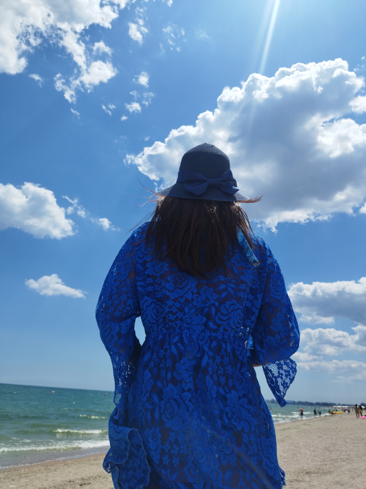 A woman wearing a blue lace dress and a matching blue hat stands on a sandy beach facing away from the camera, looking towards the sea. The sky above is a bright blue with scattered white clouds, and a sunbeam cuts across the top right of the frame. The sea is visible in the background, with gentle waves lapping at the shore. There are a few other people visible in the distance on the beach, and some buoys in the water. The scene appears to be from a sunny day at the beach, potentially in Mamaia, a resort town near Constanța, Romania, known for its long sandy beaches.