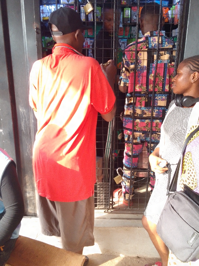 The scene depicts an outdoor commercial interaction in Nkwelle Ezunaka, Nigeria. Three individuals are prominently visible, with two others partially obscured. In the foreground, a male subject is seen from behind, wearing a bright red polo shirt, dark shorts, and a black baseball cap reversed; he appears to be reaching towards a black metal security gate. To his right, a female subject stands in profile, facing left, wearing a light patterned dress and black over-ear headphones around her neck, carrying a black shoulder bag. Behind the security gate, secured by a brass-colored padlock, a second male subject, also seen from behind, wears a distinct multicolored patterned shirt. He stands within what appears to be a stall or shop, densely packed with various electronic accessories, likely for mobile phones, displayed on shelves. A third individual is partially visible behind him, wearing dark attire. In the extreme lower-left, a portion of a person in blue and black clothing is visible next to a flat wooden surface on the light-colored ground. The scene is illuminated by strong overhead sunlight, casting defined shadows.