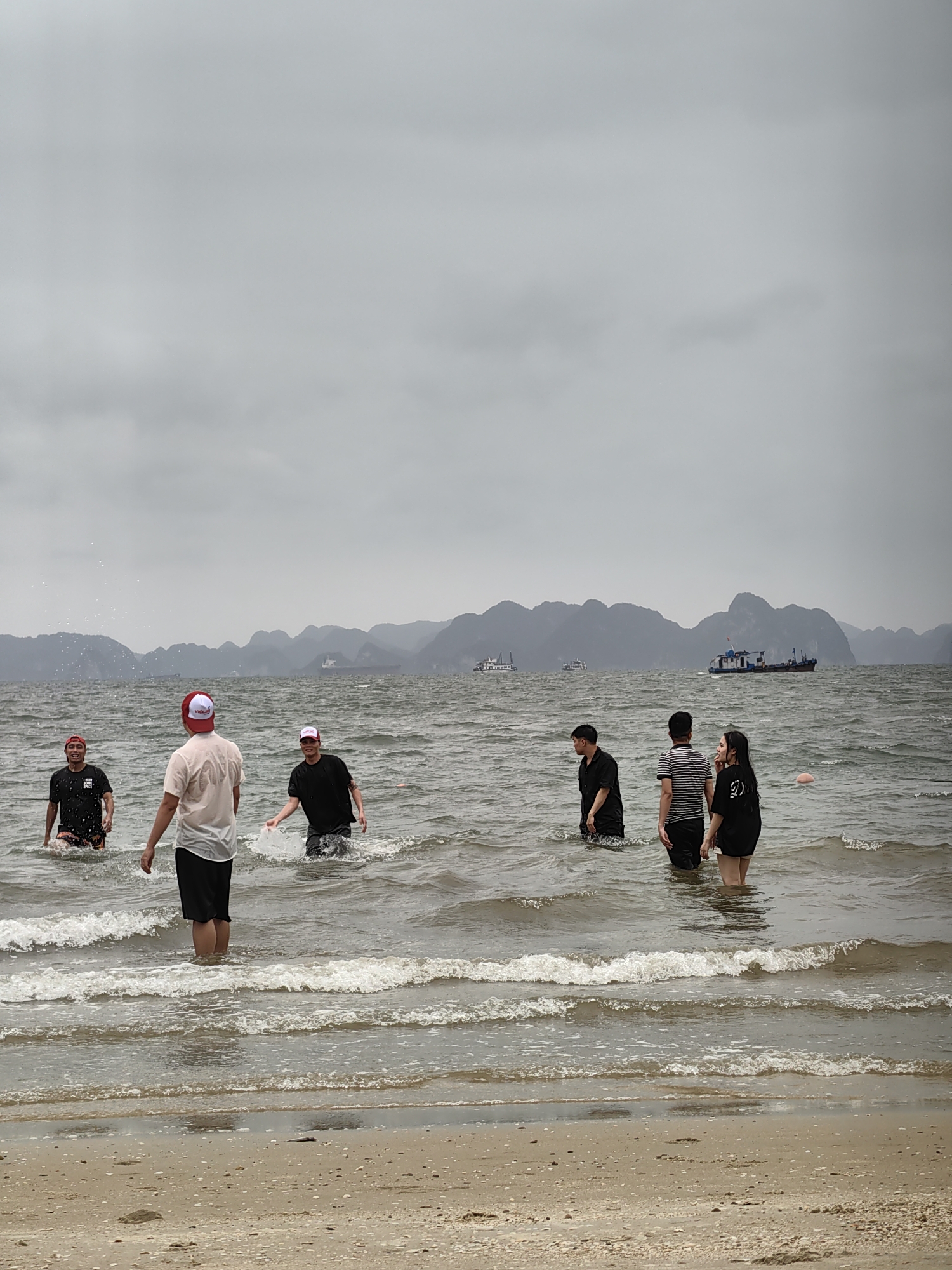 The image shows a group of six people wading in the ocean at Bai Chay Ward, Vietnam. Five are wearing dark-colored tops and dark shorts, while one is wearing a light-colored top and dark shorts. One man is wearing a hat that appears red and white. Behind the people, there is a body of water with islands in the distance, and several ships can be seen. The sky is overcast, with a gray cloudy sky. The beach in the foreground is sandy, and the waves are visible in the water.