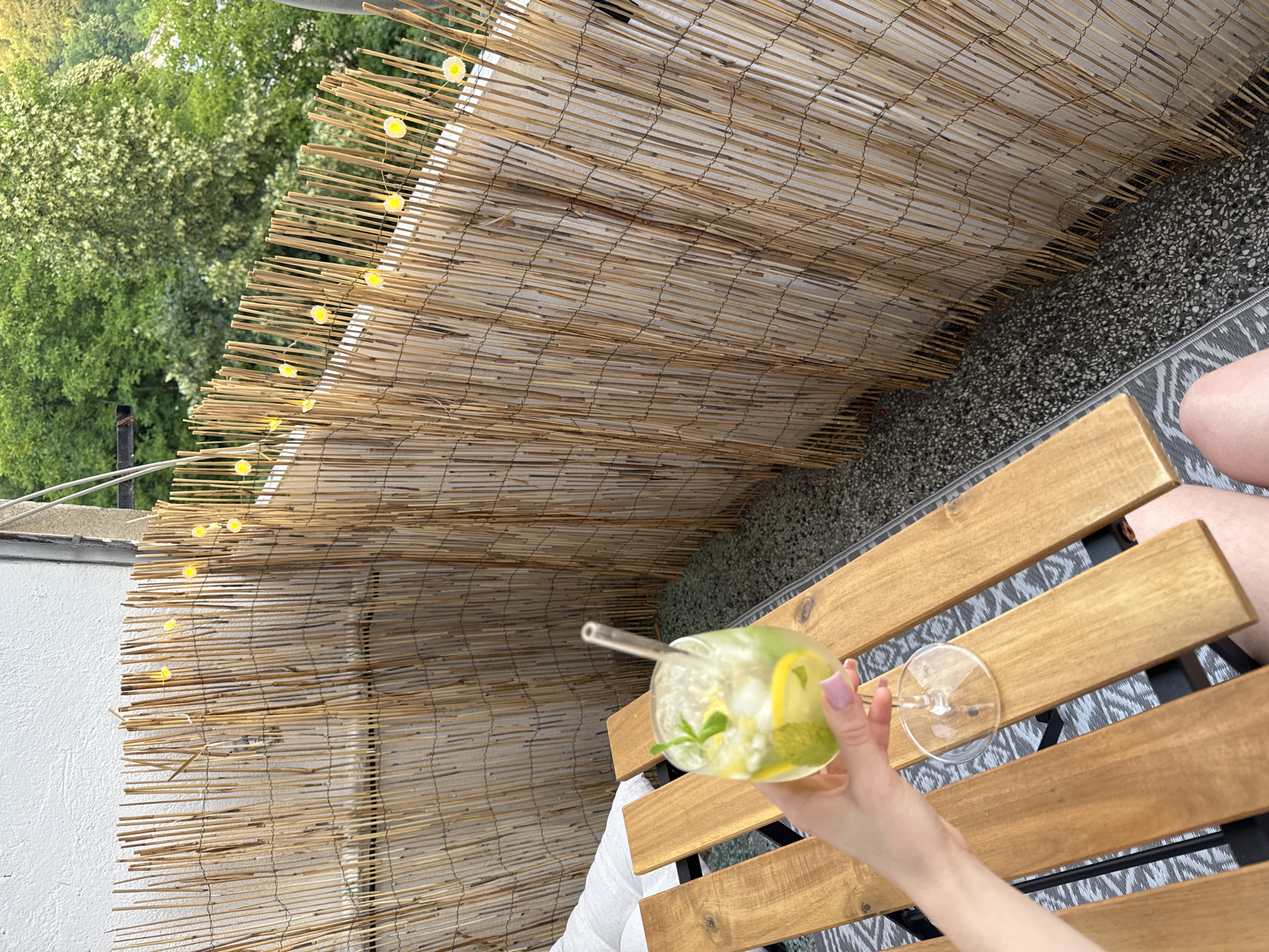 A person's hand, with painted nails, holds a clear glass filled with an iced beverage. The drink contains ice cubes, lemon slices, and mint leaves, with a straw inserted. The glass rests on the stem of itself, which is placed on a wooden surface. In the background, a privacy screen made of bundled reeds runs horizontally. Small yellow lights, resembling daisies, are strung along the top of the reed screen. The scene appears to be on a balcony or patio, with a textured floor and a wooden table. Lush green trees are visible beyond the screen, indicating an outdoor setting.