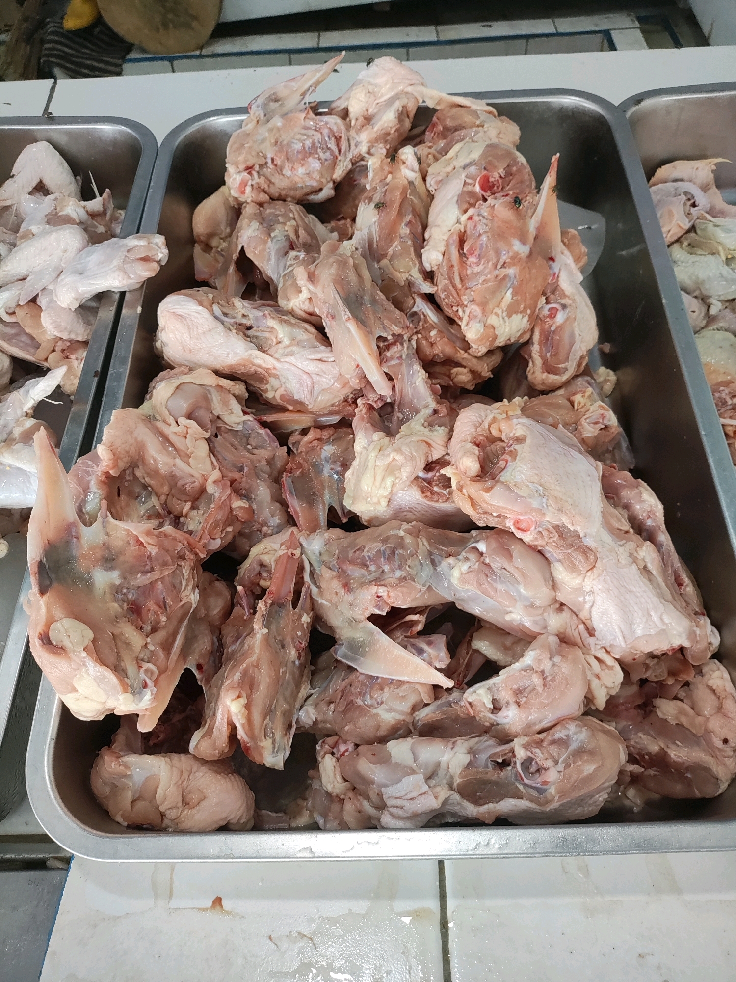 In Wanajaya, Indonesia, a close-up, overhead shot shows raw, chopped chicken pieces displayed in a large, silver metal tray. The chicken is in various cuts, including wings, legs, and torso sections, with some pieces still attached to bones. The meat is a pale pink to light brown color. The tray is positioned on a white tiled surface, with another similar tray containing chicken visible to the left. Flies are visible on some of the chicken pieces. The background shows a sliver of a striped cloth and a wooden cutting board.