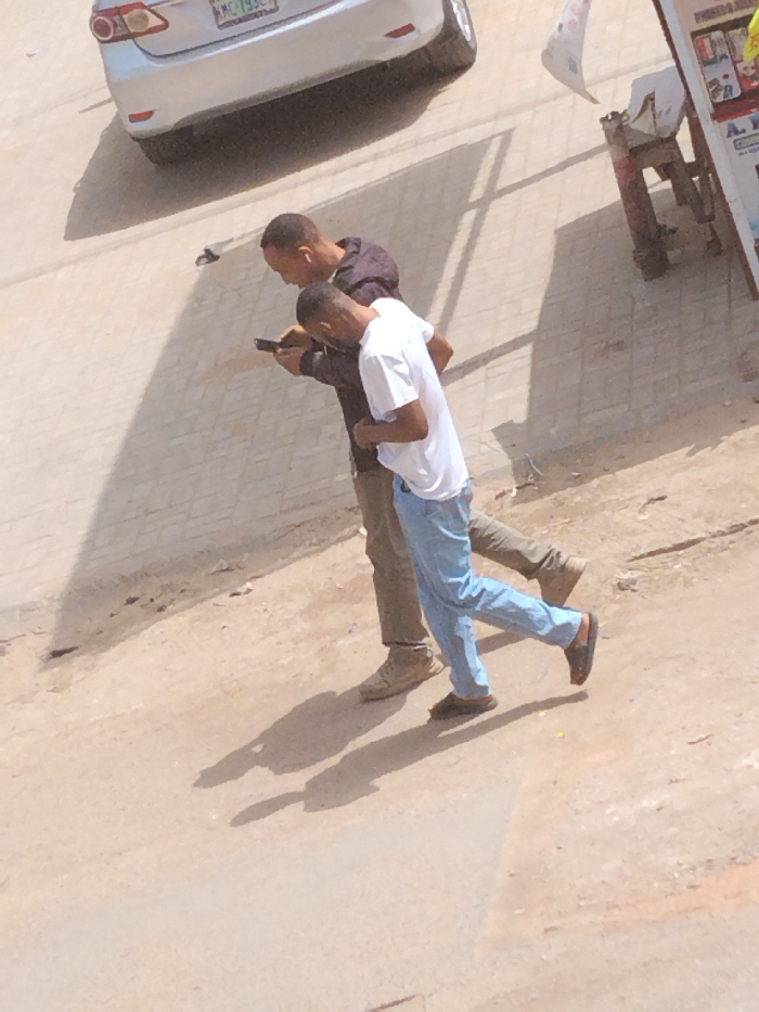 An elevated view captures two adult males walking on an unpaved, light brown surface in Fagge A, Nigeria, during daylight hours. The male in the rear wears a dark long-sleeved shirt, khaki pants, and light brown shoes, holding a smartphone in his right hand and looking at its screen. White earbuds are visible in his ears. The male in front wears a white short-sleeved t-shirt, light blue pants, and dark open-toed sandals, also directing his gaze towards the smartphone held by the other individual. Both men exhibit closely cropped dark hair and are casting distinct shadows on the ground, indicating strong overhead light. In the upper left background, a silver sedan, likely a Toyota, is partially visible on a paved surface, displaying a Nigerian license plate with green characters, partially reading "KMC 793G". To the far right, a portion of a vendor's stall or sign structure, with items displayed and a white fabric piece attached, is discernible.