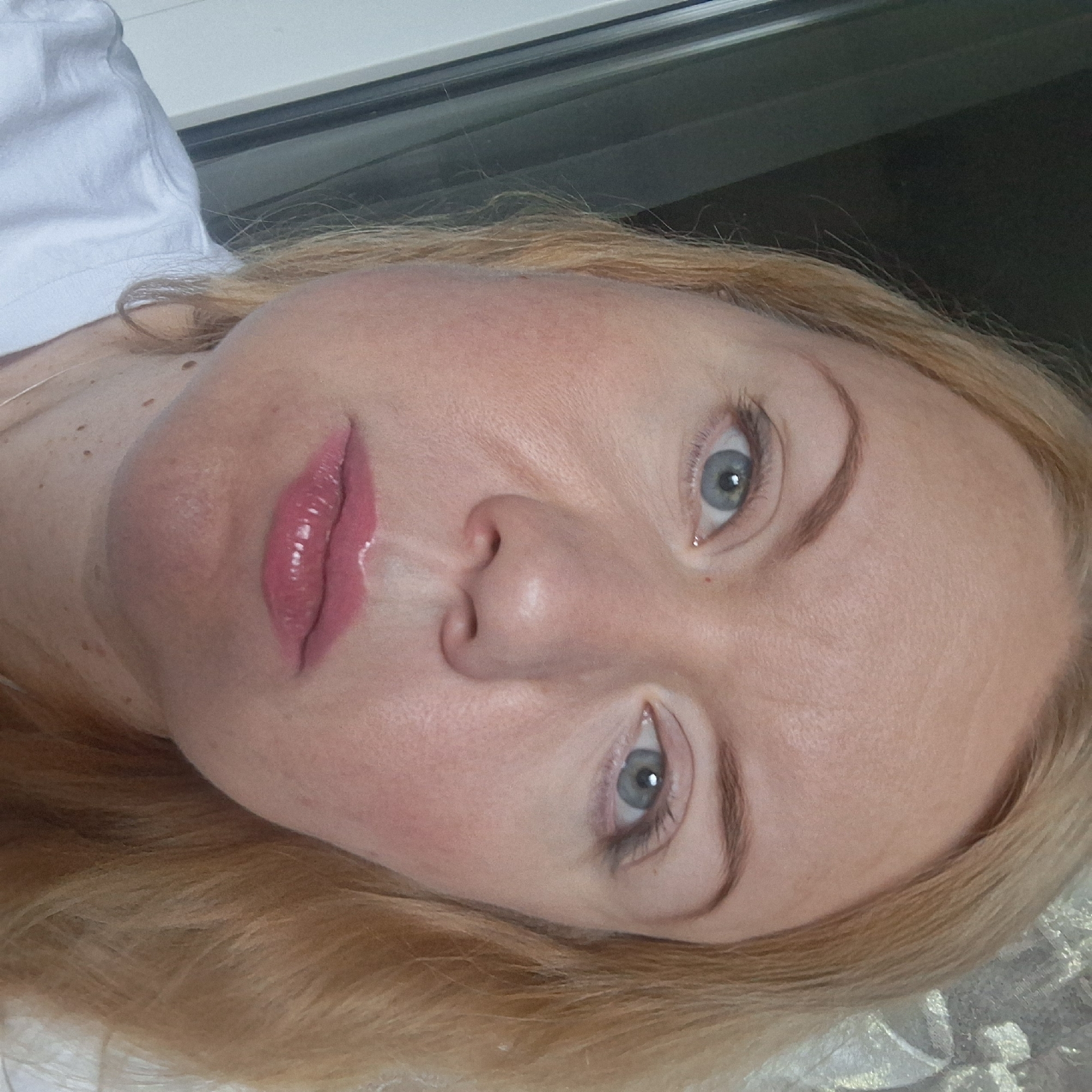 A close-up, waist-up portrait of a fair-skinned woman with blonde hair. She is wearing a white t-shirt and has bright pink lipstick on. Her eyes are a light blue and her gaze is directed slightly to the left of the camera. The background is dark on the left, with vertical lines suggesting a window frame, and a lighter, textured area on the right, possibly a curtain. The lighting is soft, casting a slight shadow on the left side of her face.