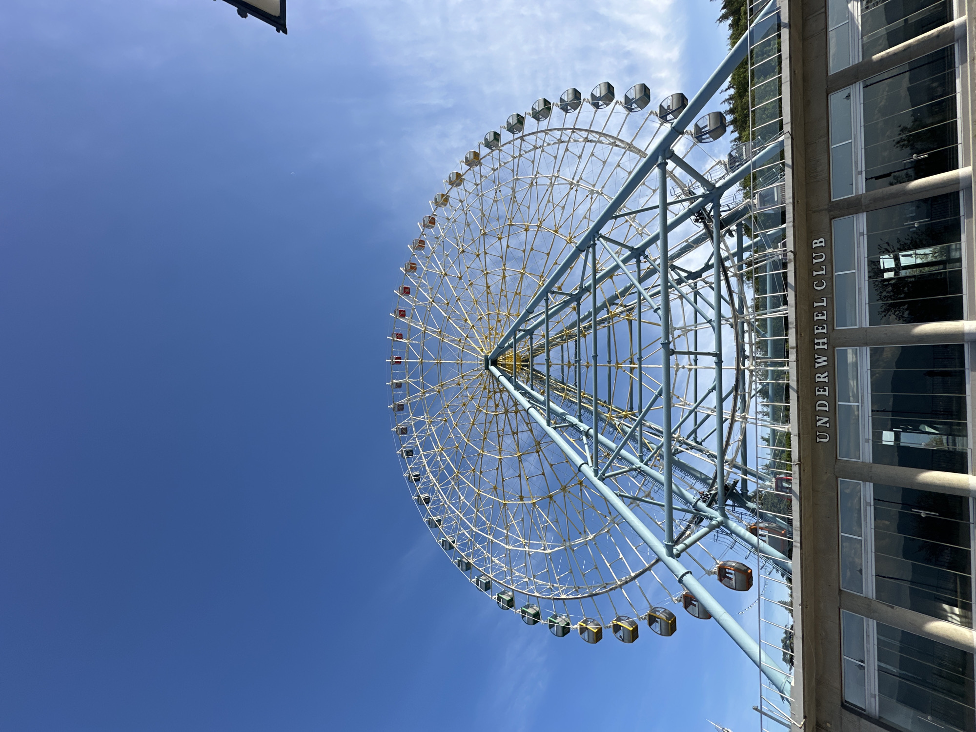 The image features a low-angle view of the Mtatsminda Ferris Wheel in Tbilisi, Georgia, set against a clear blue sky. The Ferris wheel is constructed of a blue steel frame with a network of white support cables and a central yellow core. Gondolas, some colored red, green, and brown, are visible along its circumference. Part of the "Underwheel Club" building is seen to the right of the structure, made of glass and concrete.