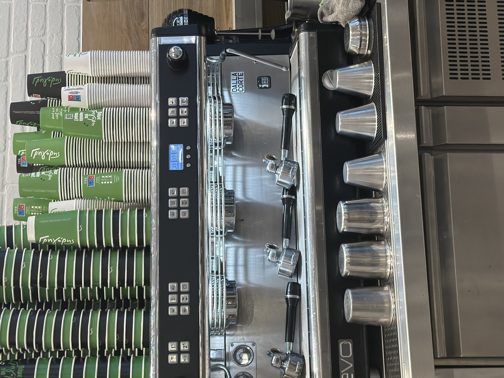 The image shows a commercial espresso machine with stacks of disposable cups on top. The machine is made of stainless steel with black accents and digital displays. Two portafilters are resting on the machine. On the counter below the machine, there are several stainless steel cups and a small metal pitcher. The scene appears to be in a cafe or coffee shop. There are no people visible in the image. The background shows a brick wall and wooden shelving. The presence of the espresso machine and coffee cups suggests a setting where coffee is prepared and served. The location in the city of Municipal Unit of Mantineia, Greece, is not evident from the visual information in the image alone.