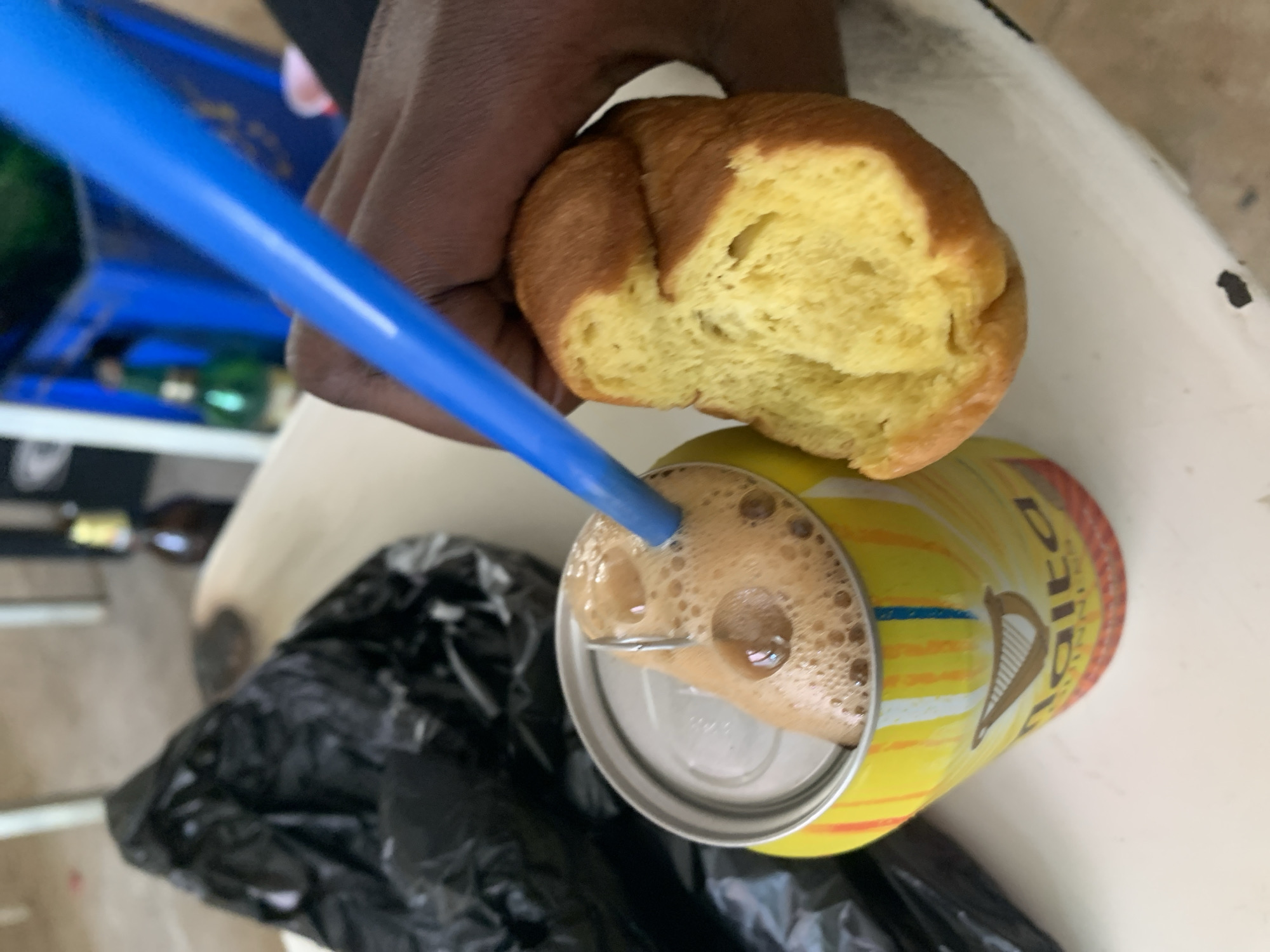 In New Stadium, Nigeria, a person's hand is holding a piece of bread over a can of Malta Guinness. The can is yellow with red and blue stripes and features the Guinness harp logo. A blue straw is inserted into the can, and the drink is frothy. A black plastic bag is visible in the background, as are some green bottles and blue crates. The scene appears to be outdoors, possibly on a table.